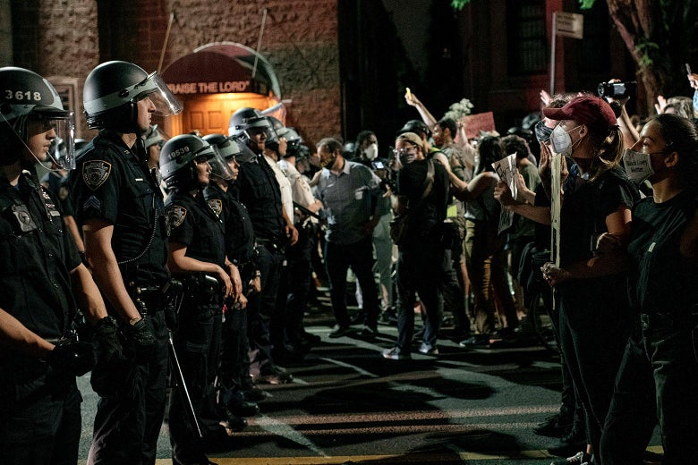 After dark, a line of NYPD officers with batons stare down a group of protesters.