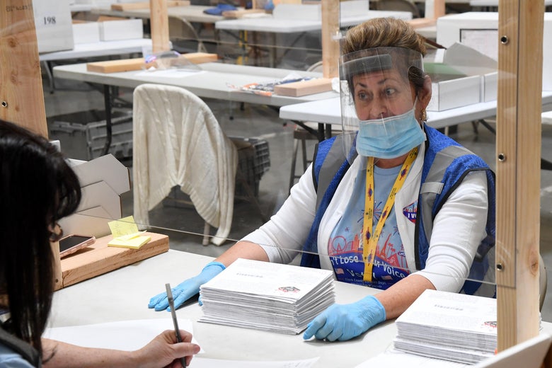 Thompson wears a face shield, mask, and gloves and sits behind a clear screen across from someone filling out a form.