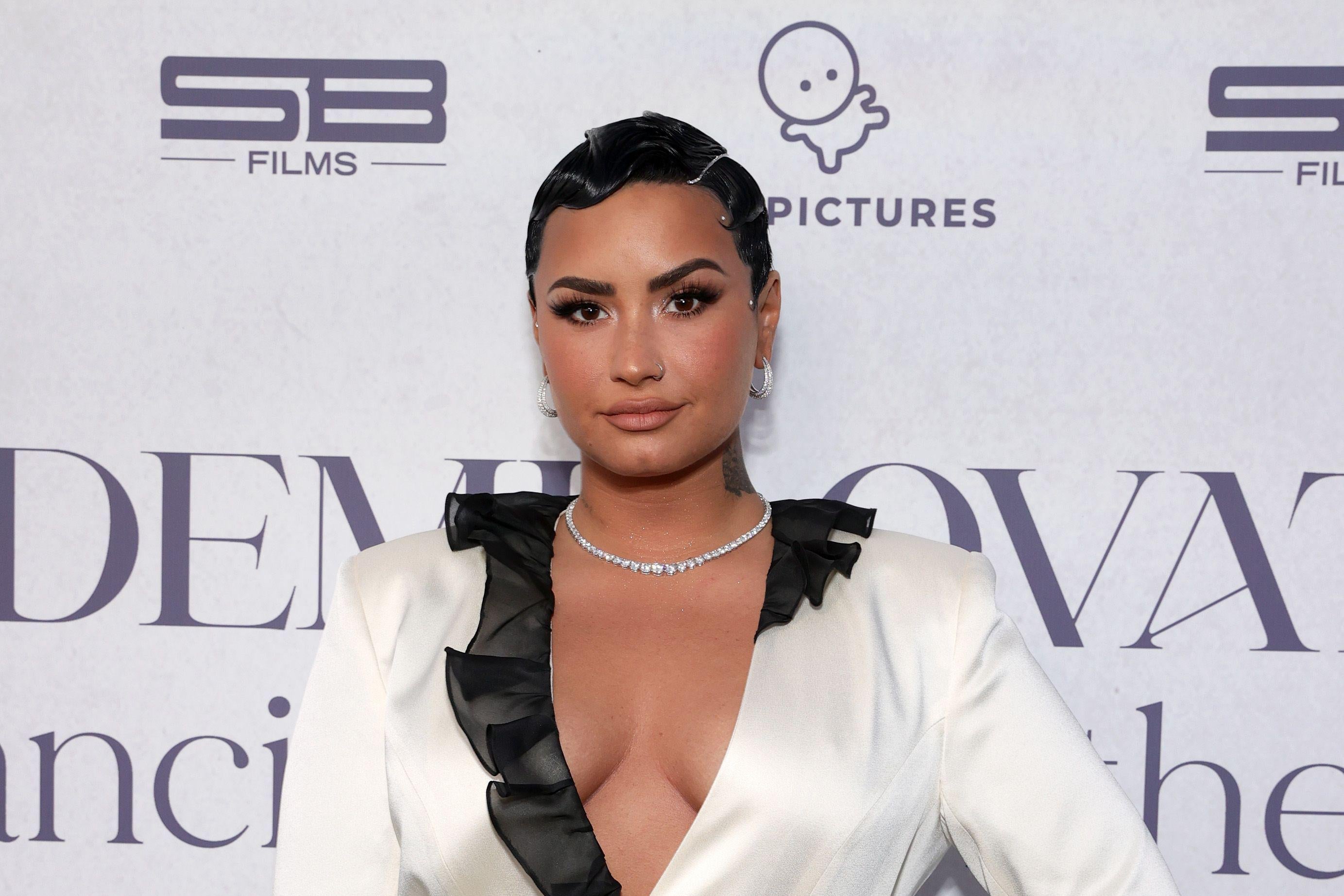 Lovato on the red carpet sporting a black wavy pixie cut and a white dress with a plunging neckline, black cuffs, and a black ruffled collar.