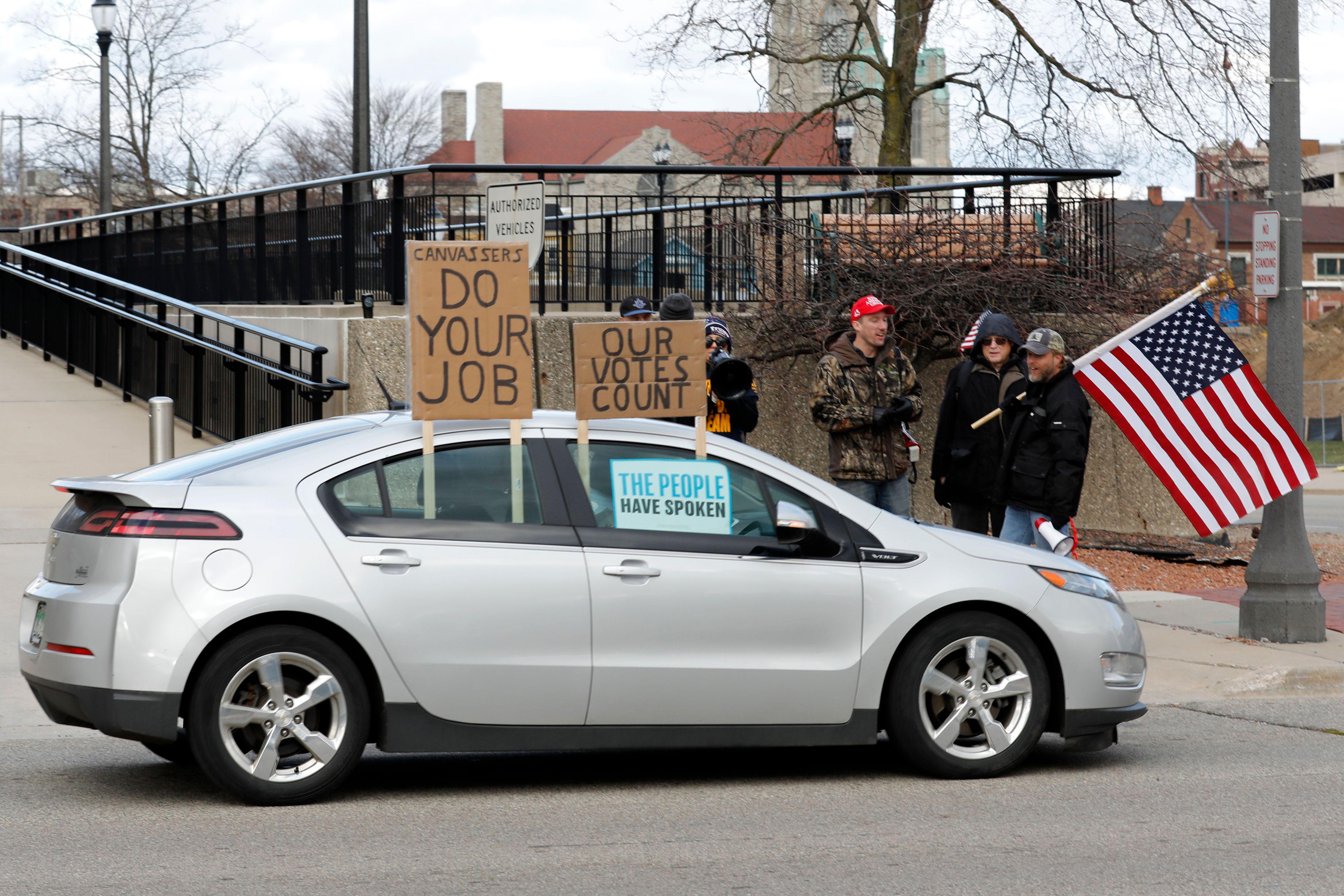 A car with signs on the window that say "DO YOUR JOB," "OUR VOTES COUNT," and "THE PEOPLE HAVE SPOKEN" drives by a group of protesters, one of whom is holding an American flag