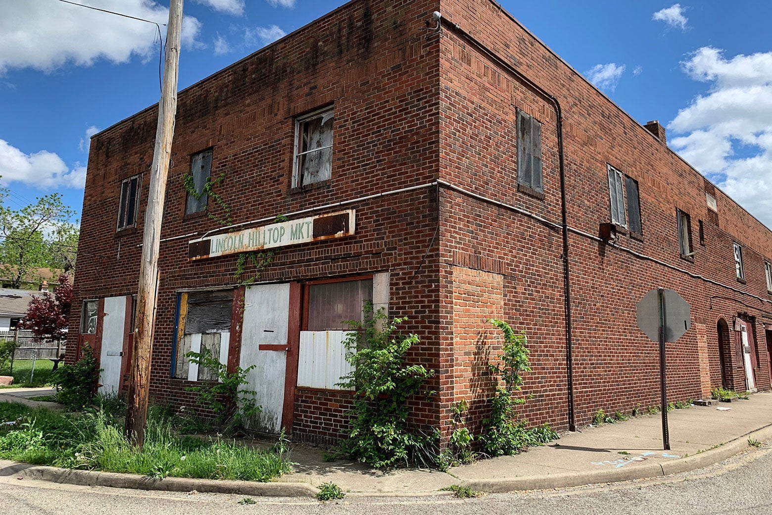 A brick two-story building with boarded-up windows on a street corner. A rusted sign says "Lincoln Hilltop Mkt."