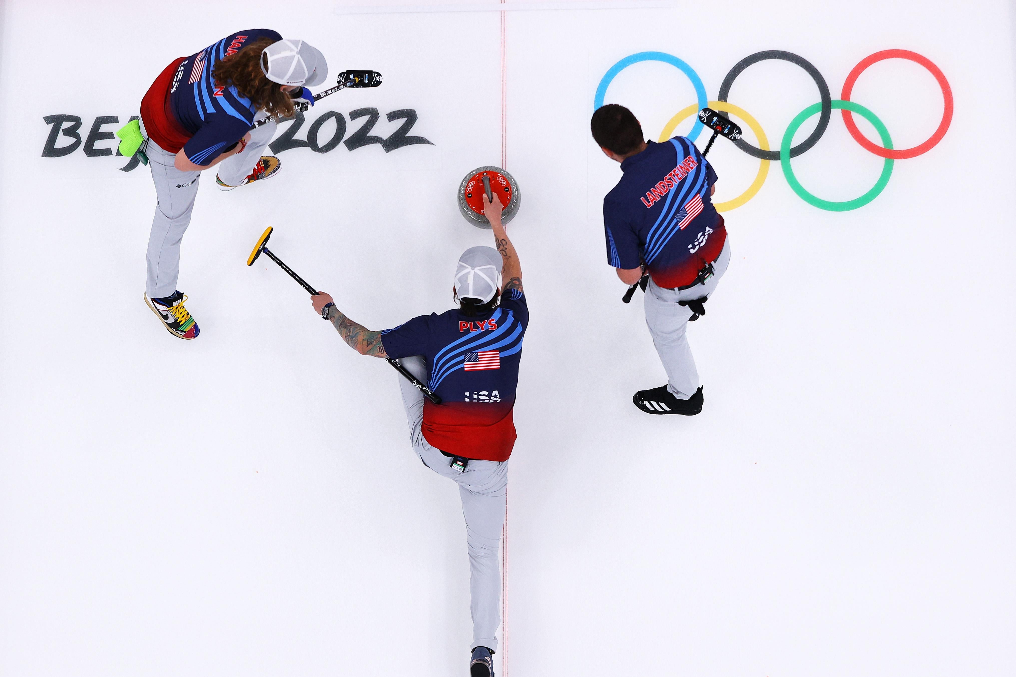 Three dudes in USA uniforms curling, the middle guy pushing to rock and the others brooming away, seen from above, with Olympics rings and Beijing 2022 logos on the ice