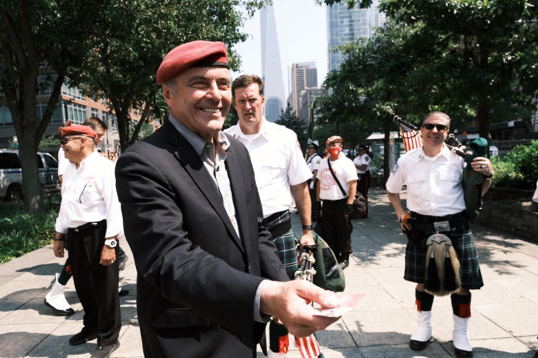 Sliwa smiles in a suit and red beret, holding out what appears to be a business card. Police and other first responders including two men in kilts stand nearby outside on a sunny day.