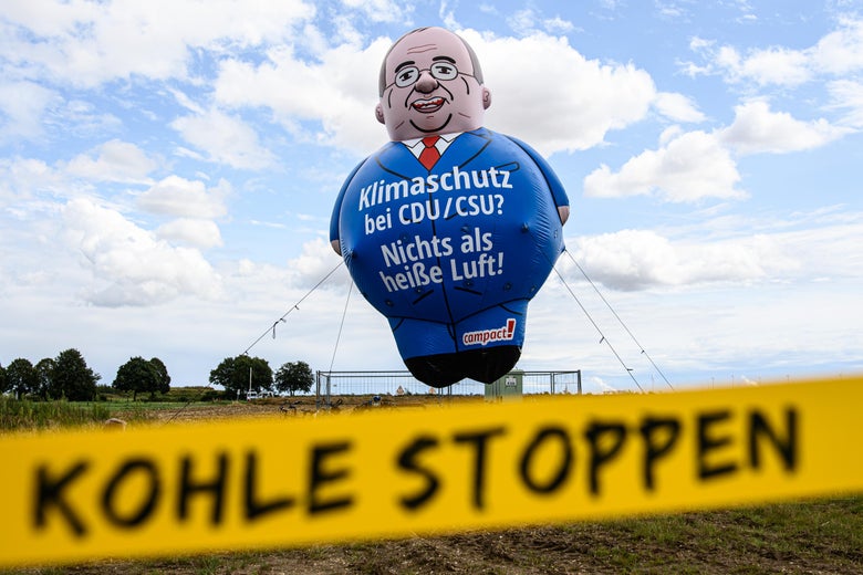 A balloon in the shape of a smiling man wearing a blue suit and glasses floats above a field with a banner in the foreground that says "Kohle Stoppen" or "Stop Coal"