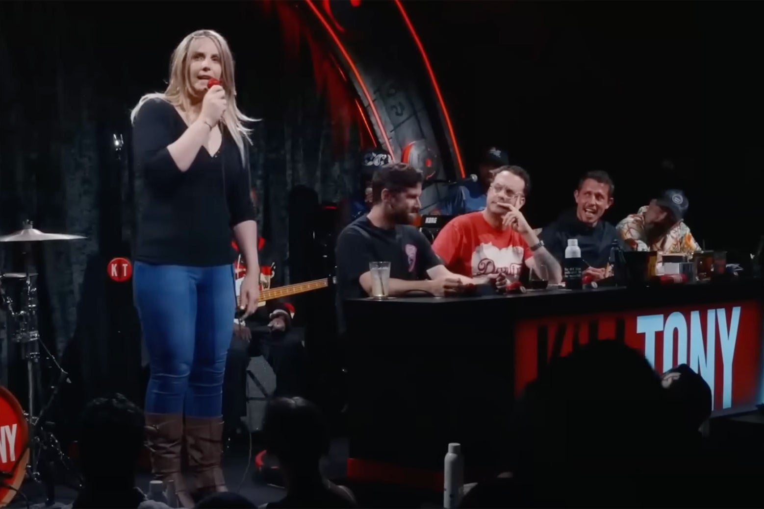 A woman in jeans and cowboy boots stands on stage next to a panel of four men behind a desk.