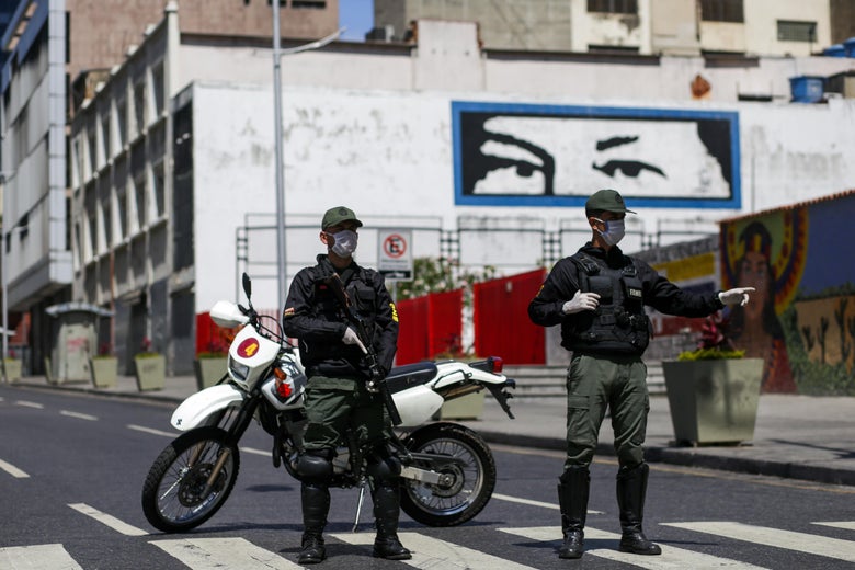 Troops carrying weapons and wearing facemasks stand by a roadblock in front of a mural of Hugo Chavez.