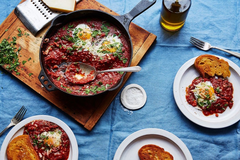 Cast iron pan filled with tomato-and-egg dish, surrounded by plates with servings of the same dish, laid out on a blue tablecloth.
