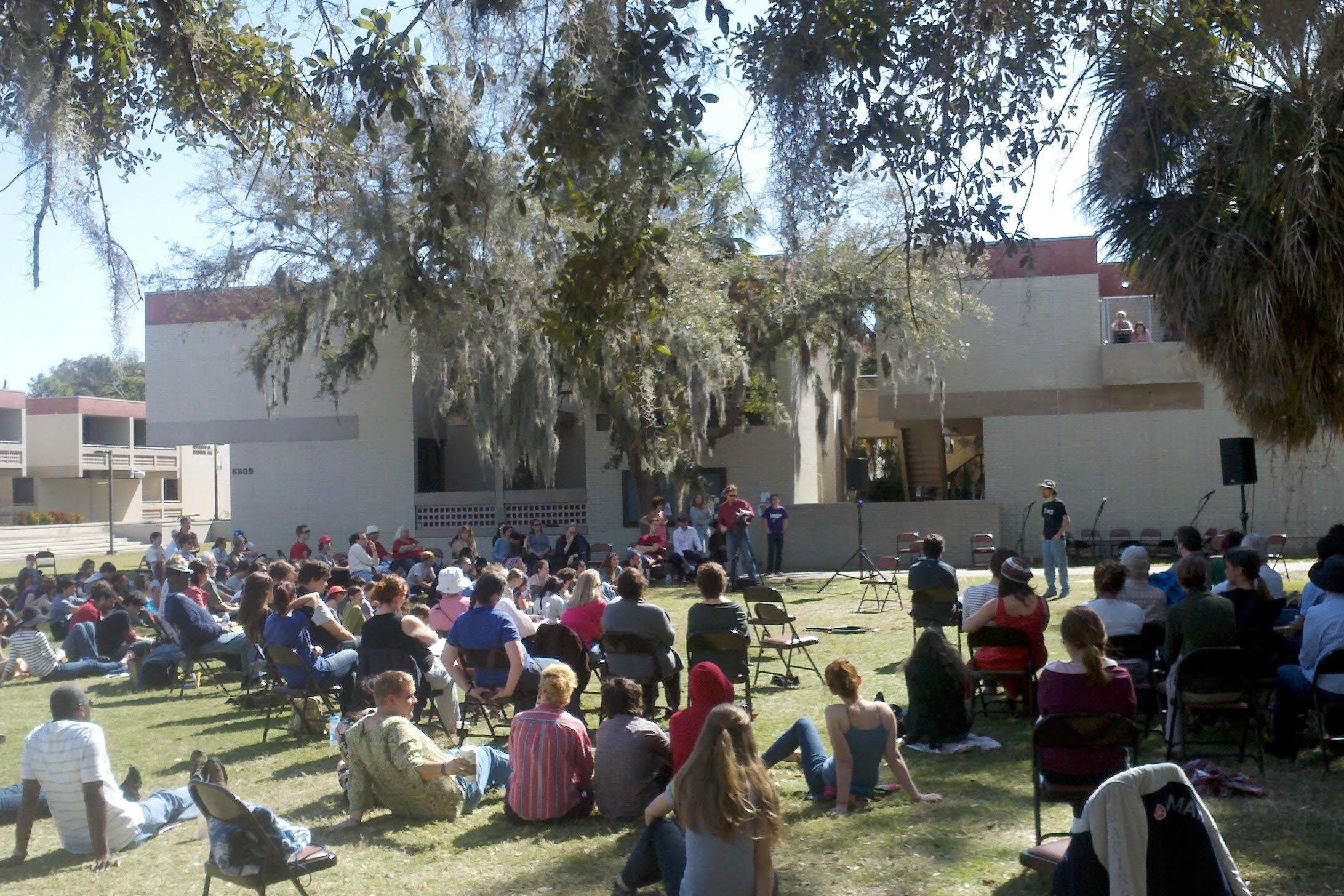 On a lawn with palm trees and oak trees with Spanish moss, a crowd of students sit on the ground or in folding chairs.