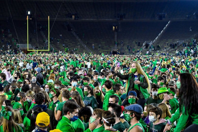 Hundreds (possibly thousands) of young fans wearing green, most wearing masks, are seen against a backdrop of empty bleachers.