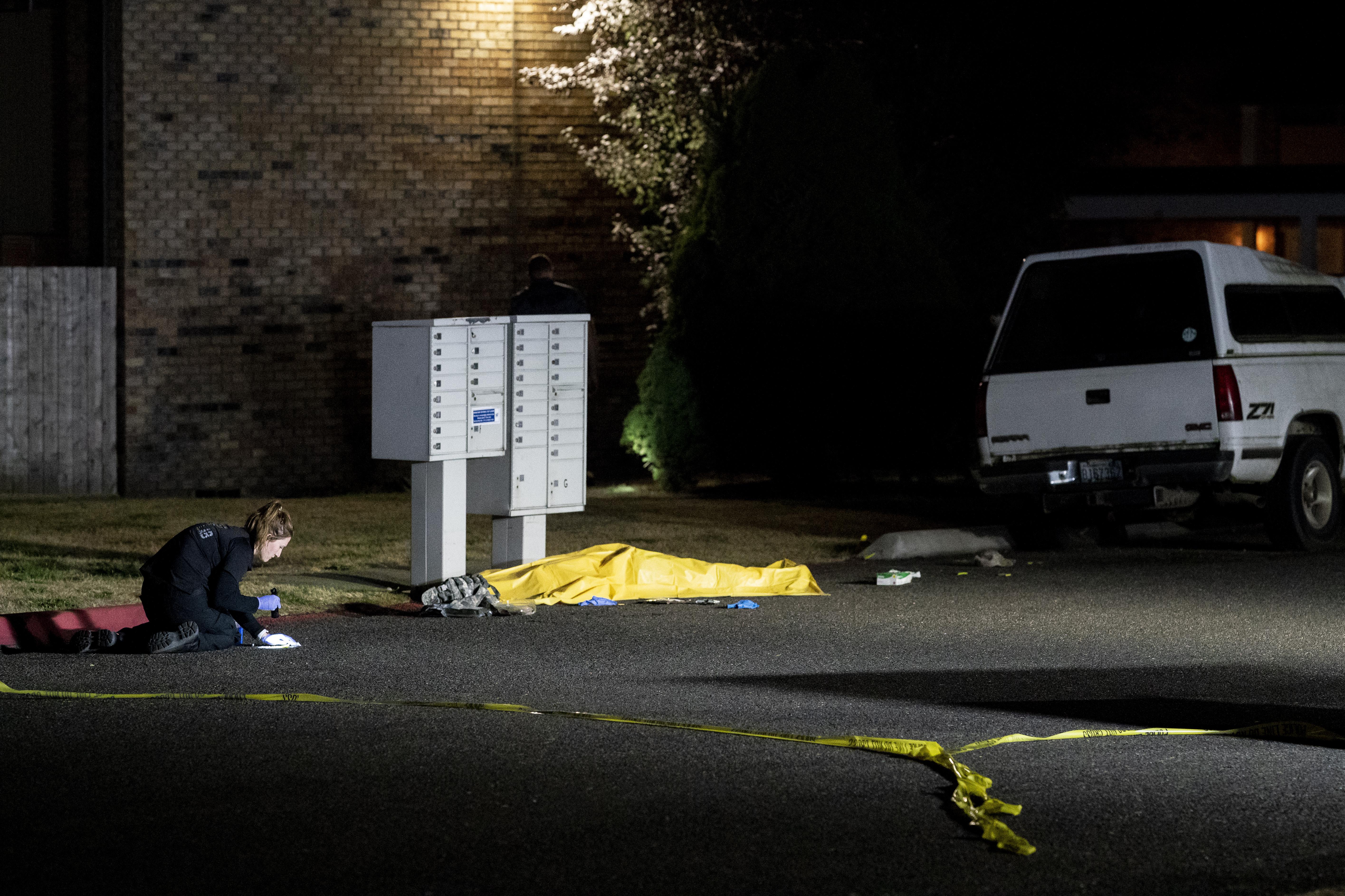 An investigator, kneeling, inspects something on the ground next to the dead body, which is covered by a tarp underneath a mailbox
