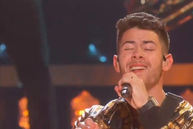 Nick Jonas sings into a Grammy microphone with his eyes closed and a piece of food sticking out of his teeth.