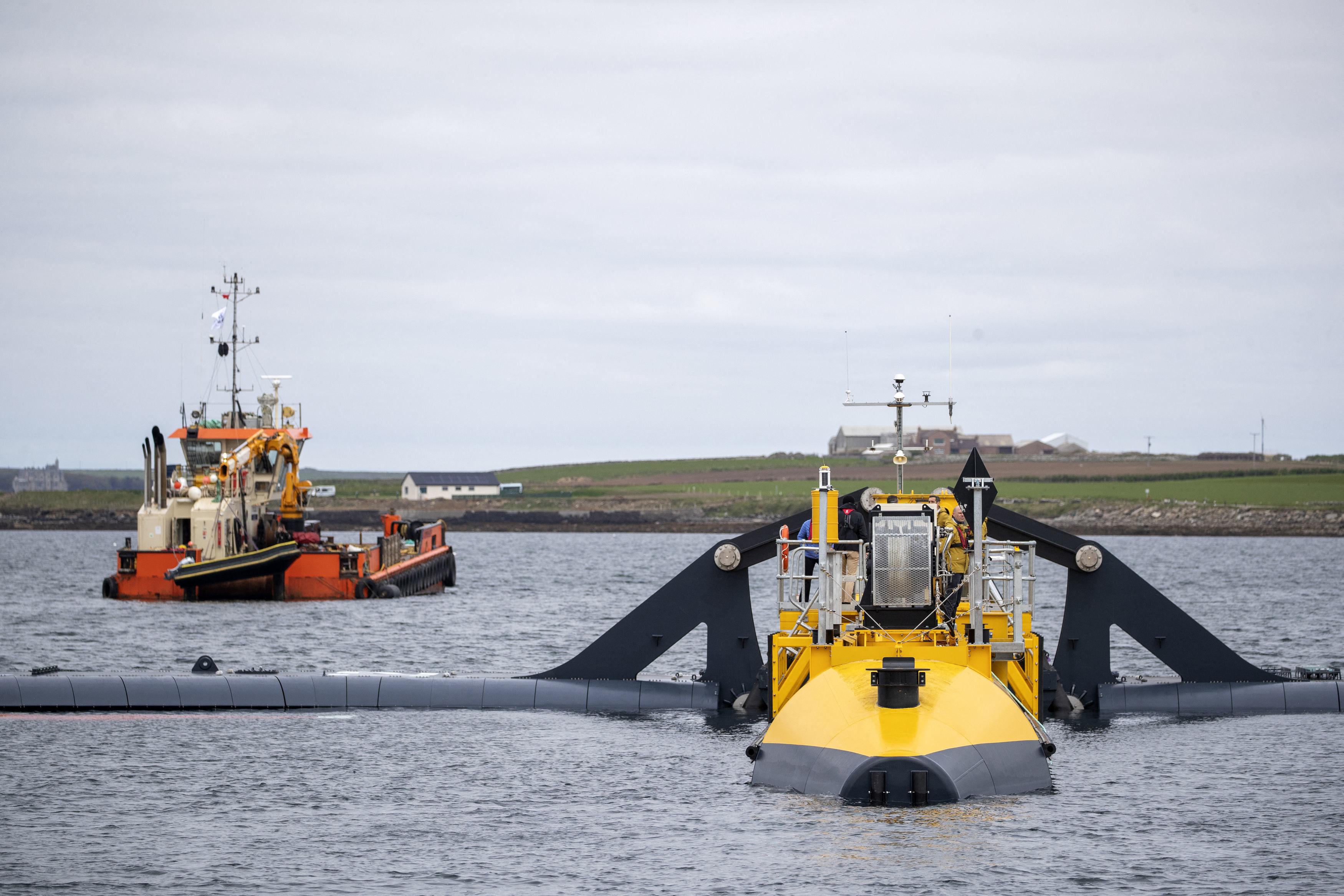 A boat and a tidal generator sit in the water with land in the background.