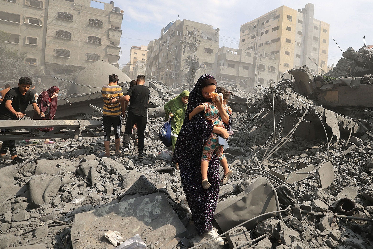A woman holding a child walks over rubble from a concrete building. Several people are climbing over the rubble in the background.