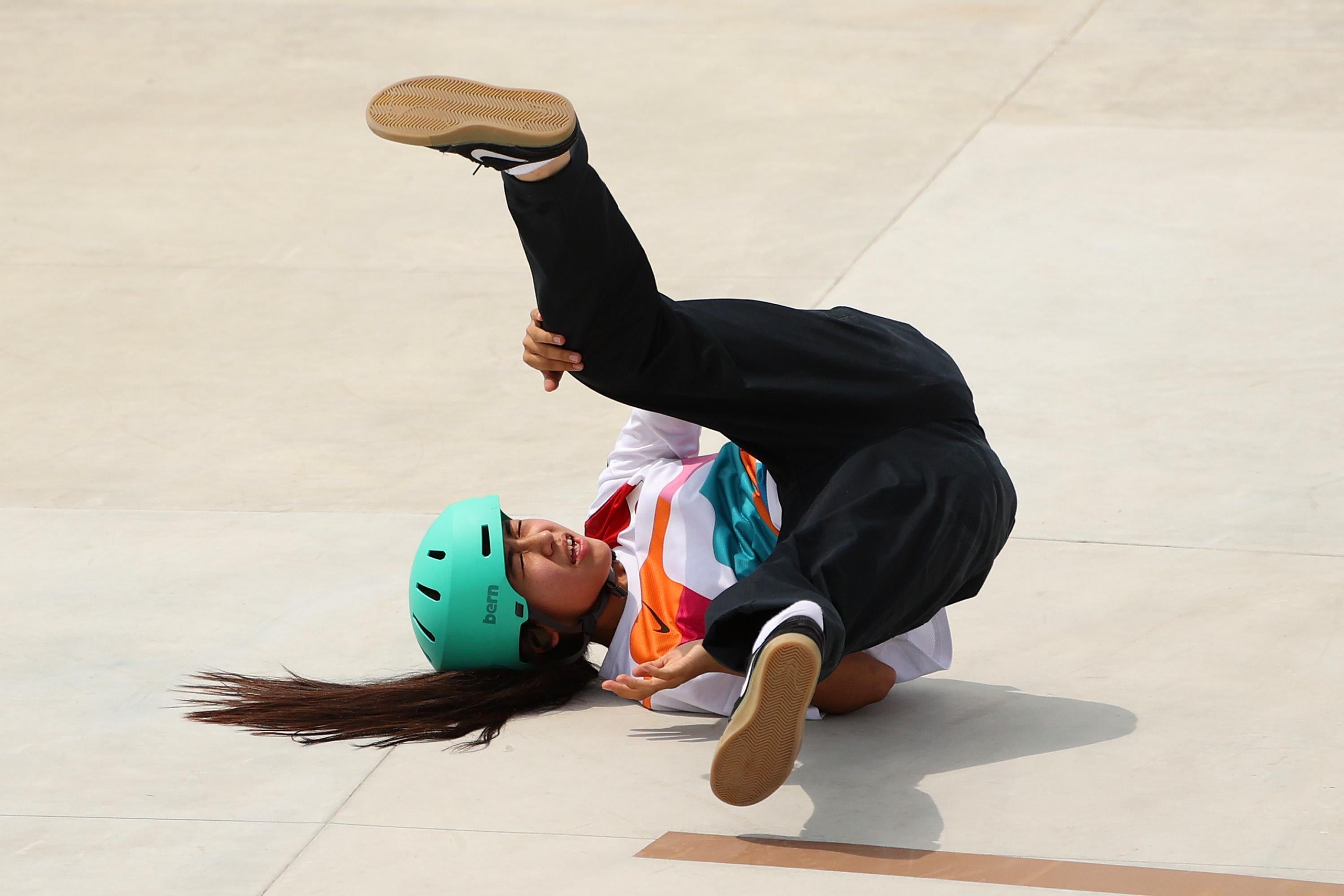 A girl in a teal helmet and long ponytail falling on her back, legs in the air, on concrete