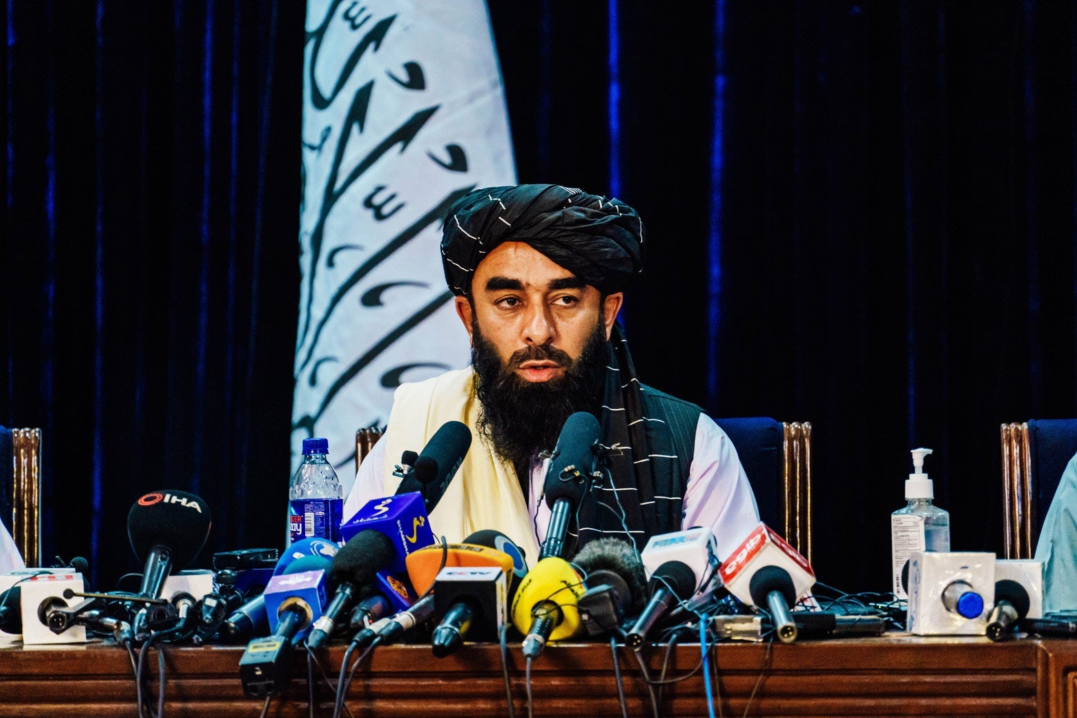 A man in with a beard and his head covered sits behind a row of microphones.