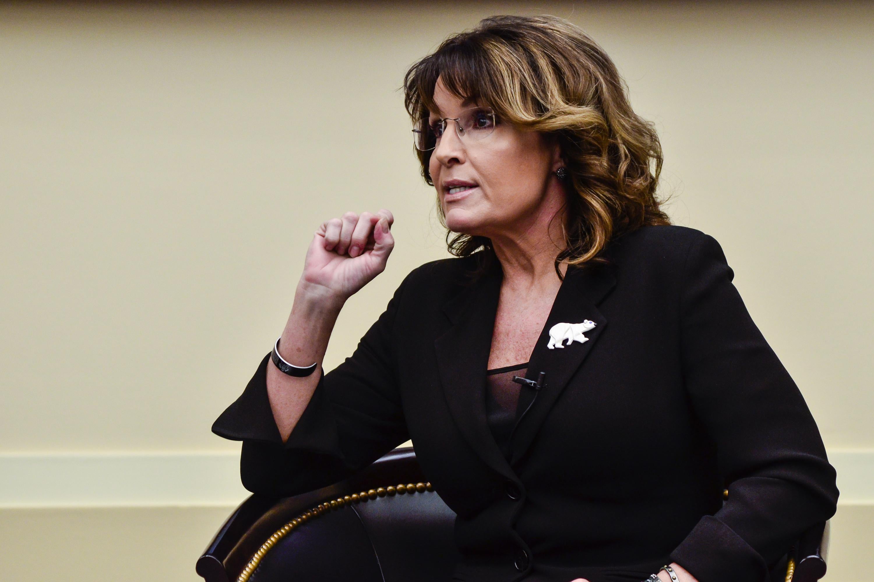 Sarah Palin sits in a chair, speaking. She wears a pin with a bear on it.