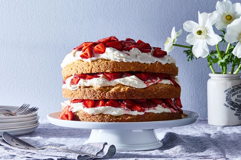 A three-layer vanilla cake doused in whipped cream and fresh strawberries sits on a white cake stand.