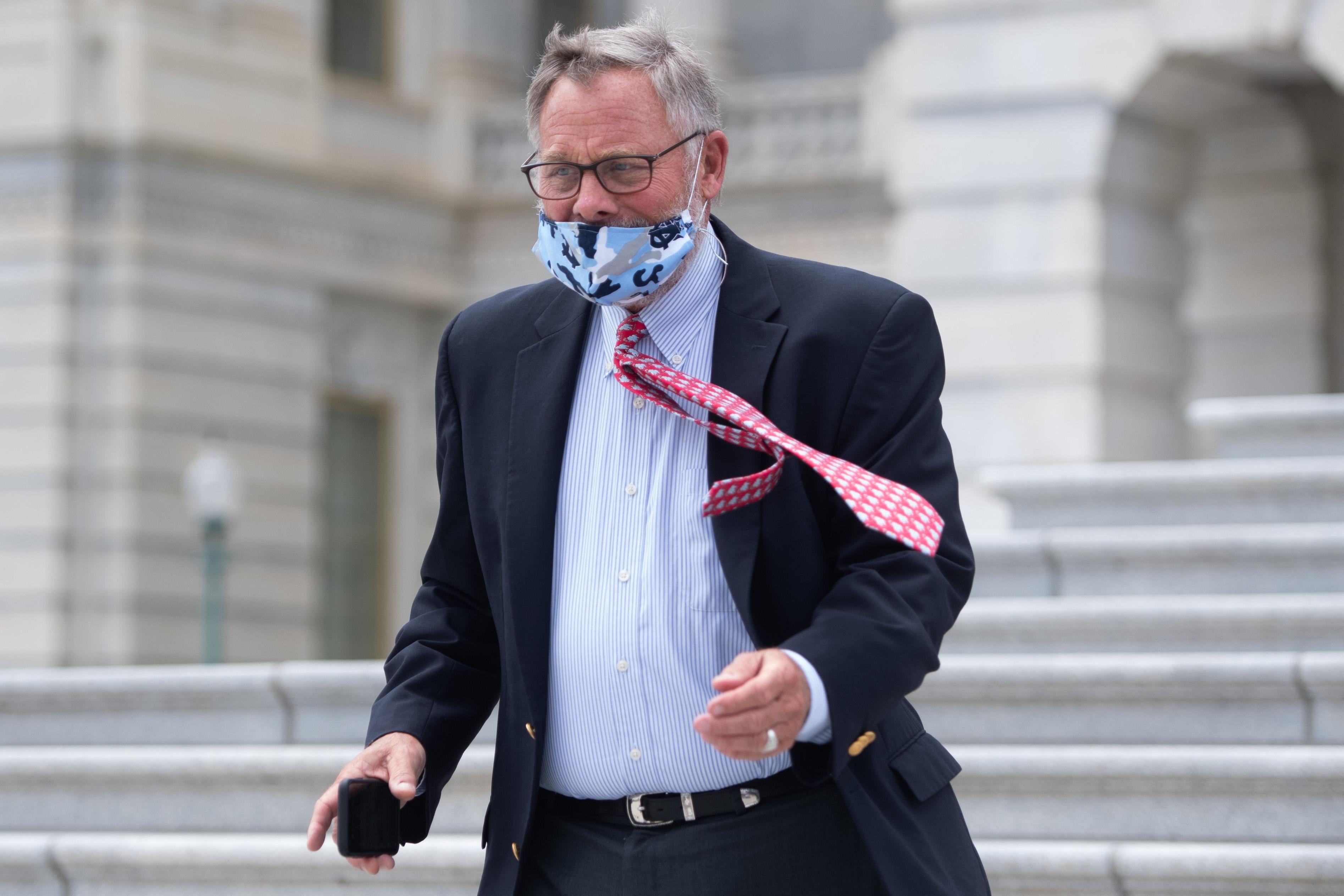 Richard Burr, wearing a face mask and carrying his cellphone, walks down stairs as wind blows his tie to the side.