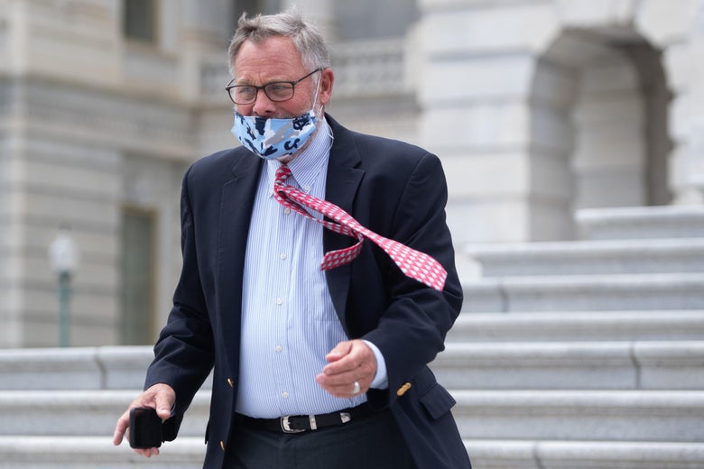 Richard Burr, wearing a face mask and carrying his cellphone, walks down stairs as wind blows his tie to the side.