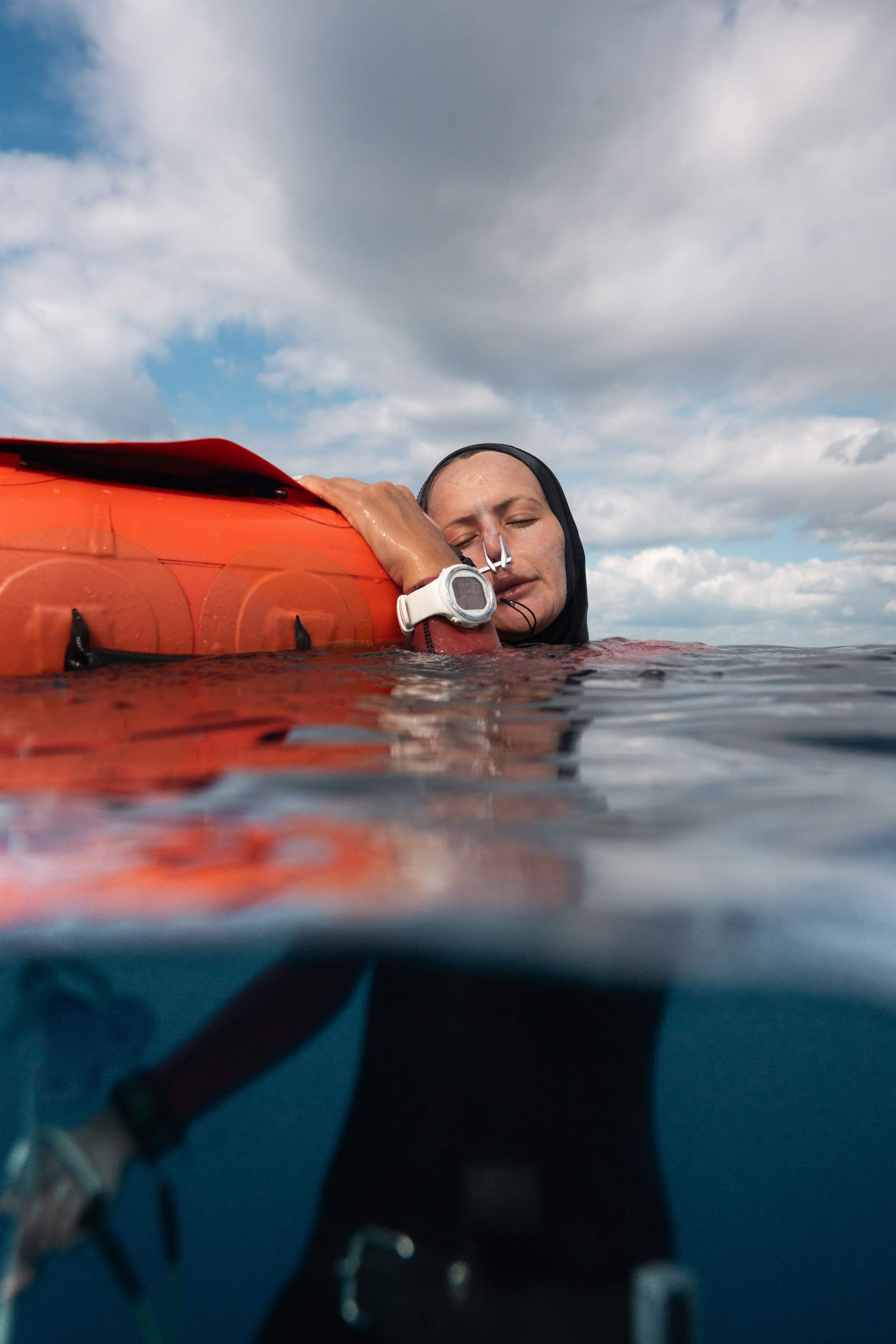 A woman in a wetsuit leans her head on an orange raft she is hanging onto.
