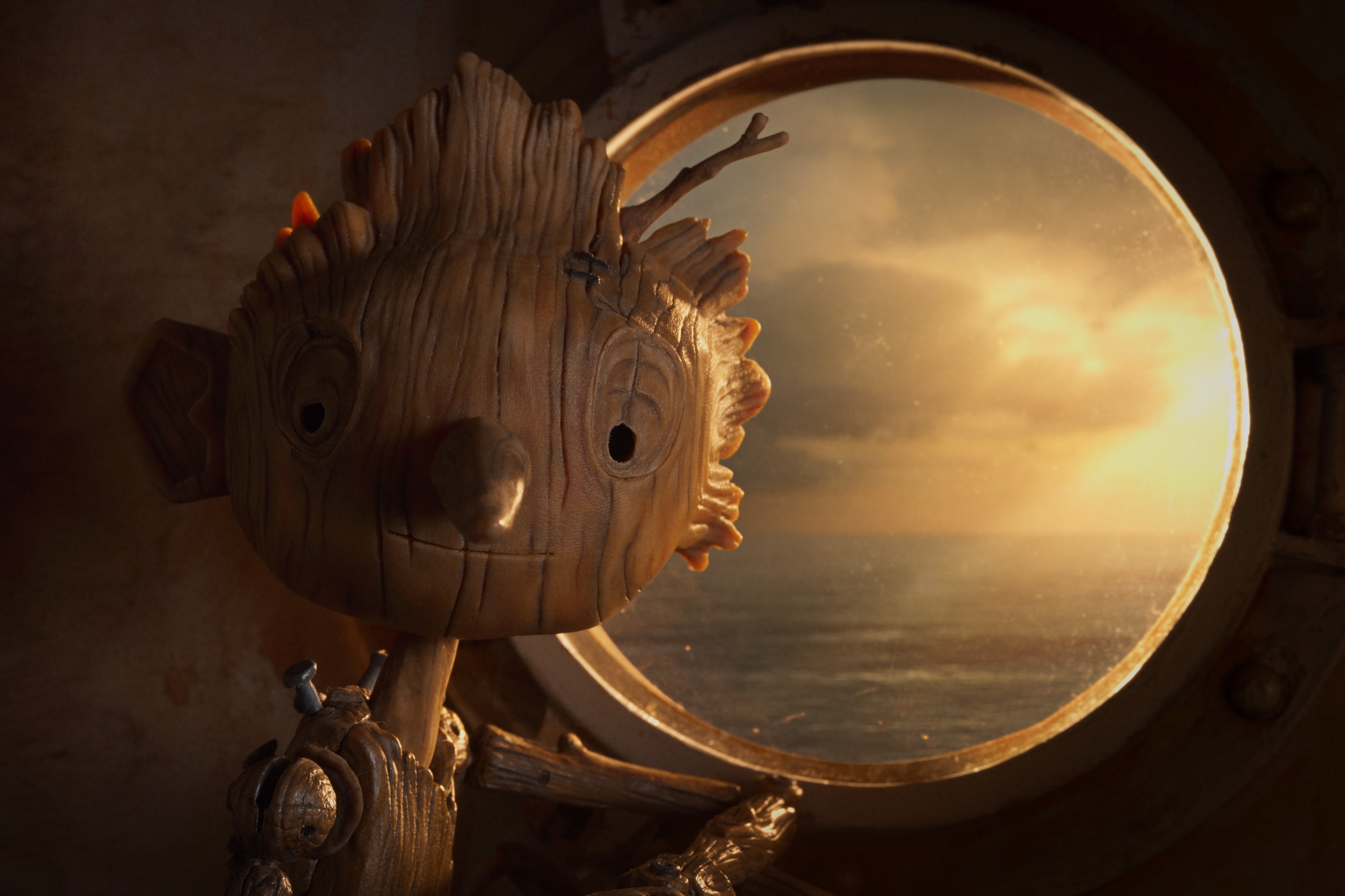 A wooden boy stares into the camera, a faint smile on his face, as golden light filters through a porthole window behind him