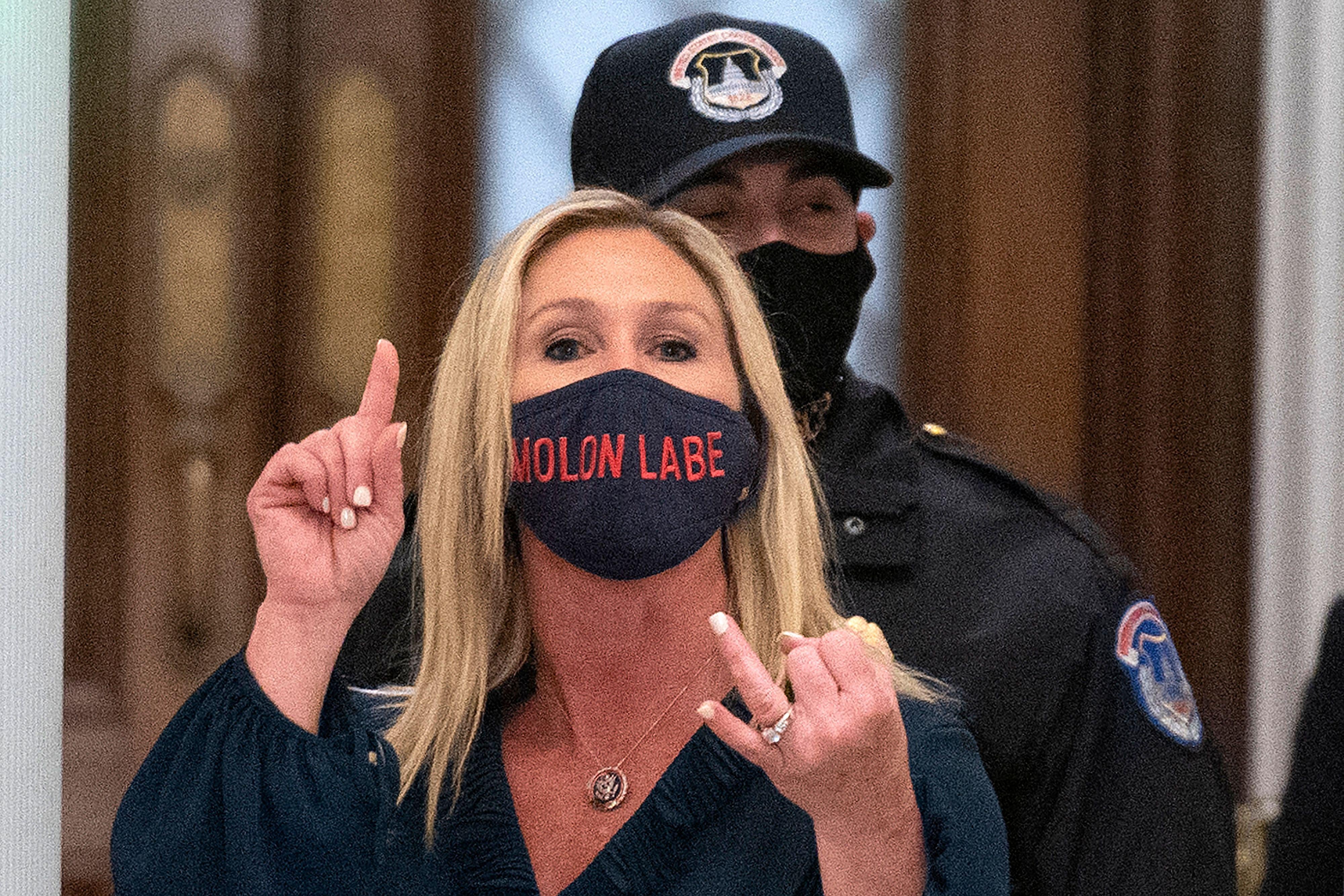 Greene, standing in front of a metal detector, gestures with her hands and wears a mask that says "Molon Labe," which means "come and take them" in Greek.