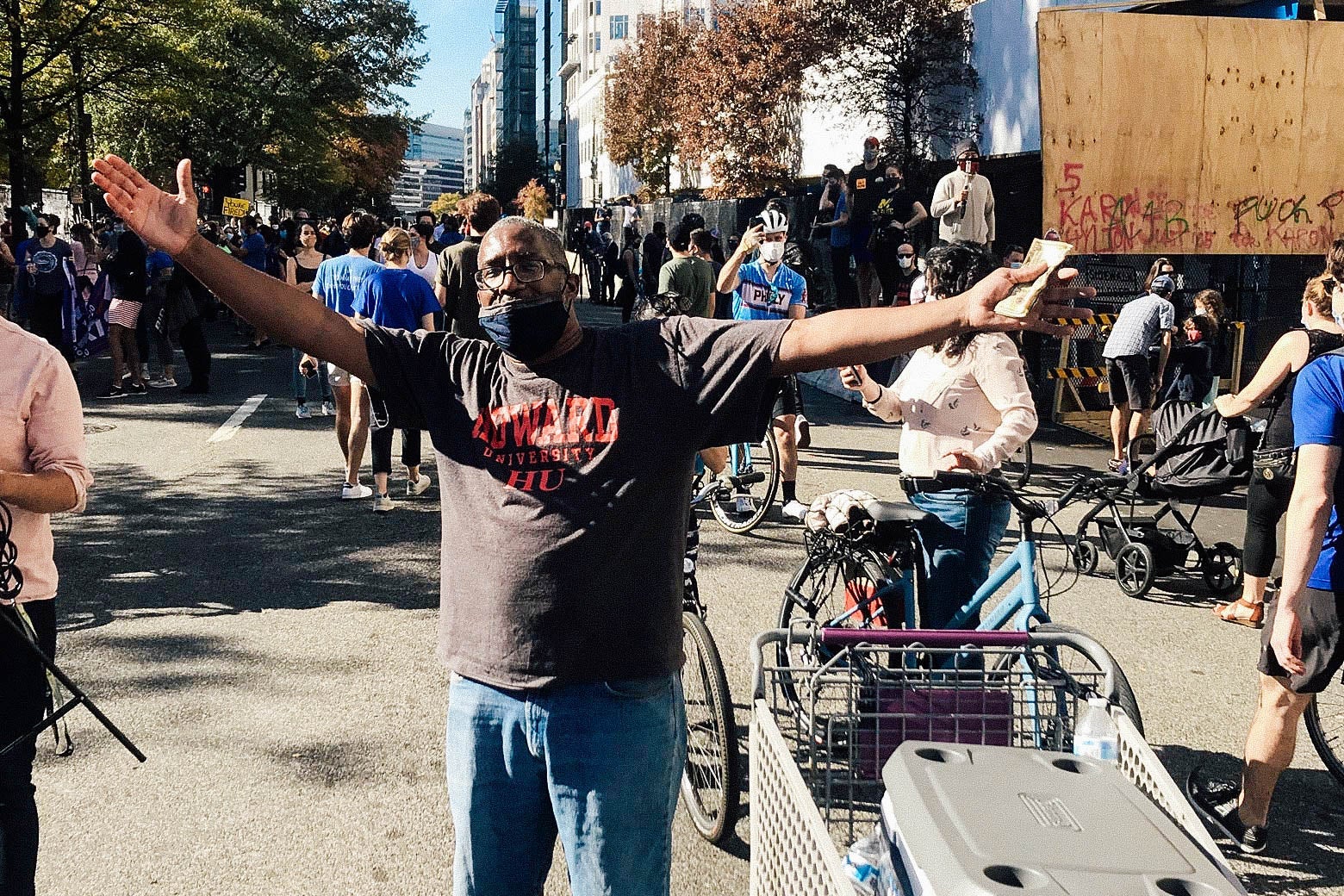 A man stands with arms outstretched next to a cooler full of water. In the background, a gathered crowd.