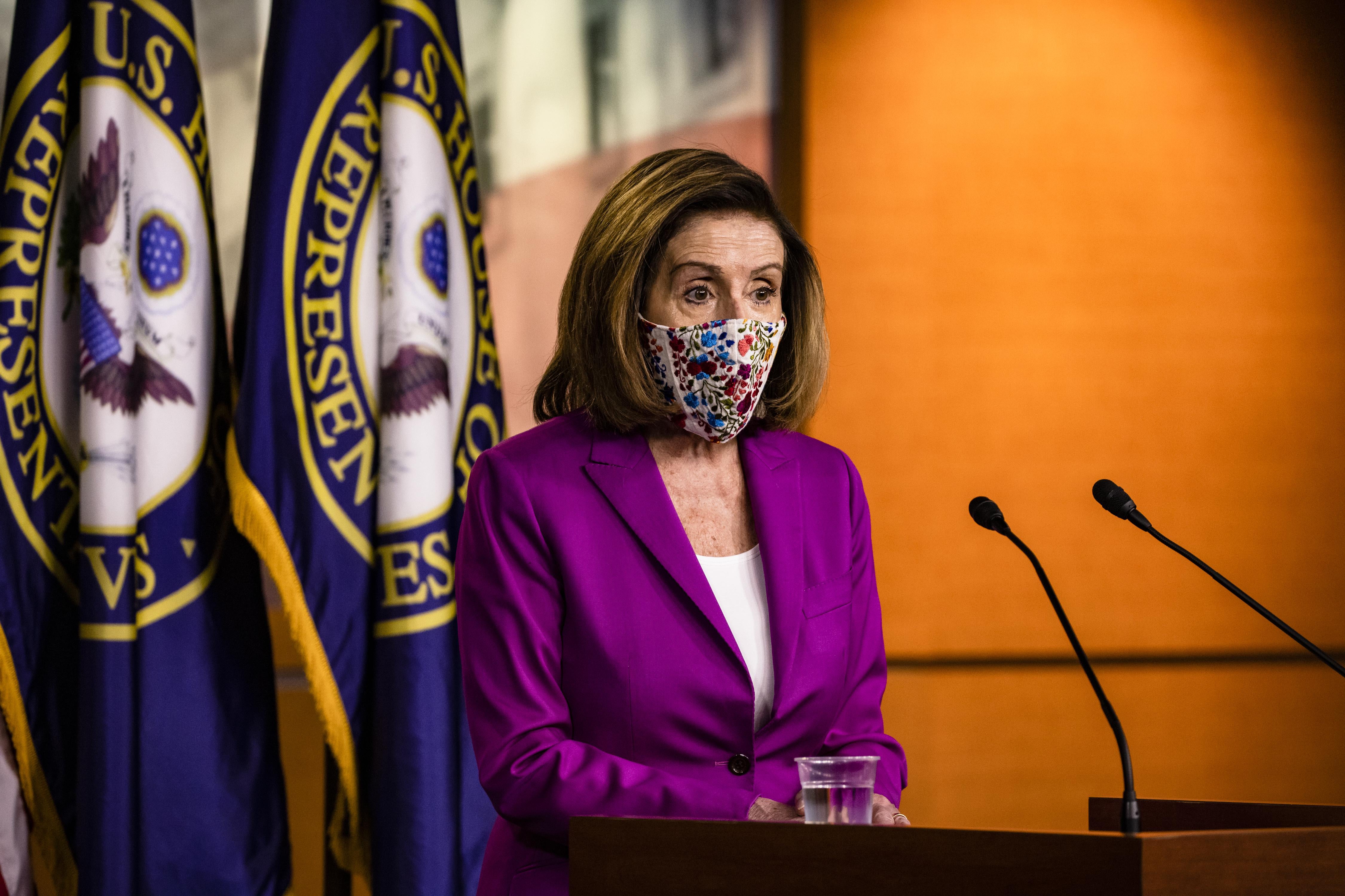 Pelosi speaks into two microphones while standing in front of two House of Representatives flags.