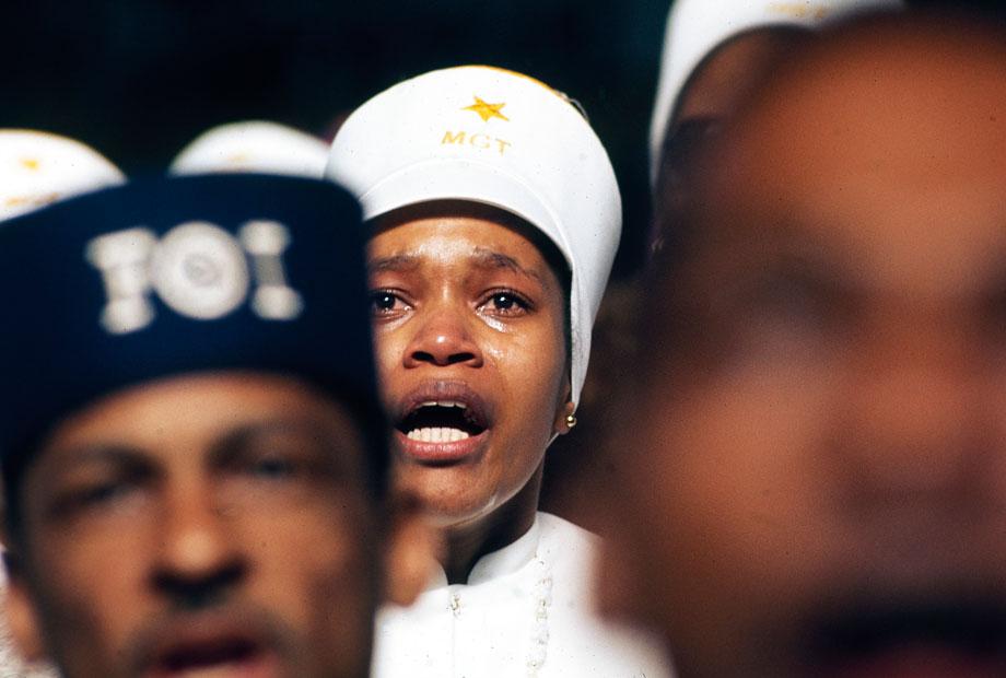Religious fervor is mirrored on the face of a Black Muslim woman, one of some 10,000 listening to Elijah Muhammad deliver his annual Savior’s Day message in Chicago. The city is headquarters for the Black Muslims. Their $75 million Empire includes a mosque, newspaper, university, restaurants, real estate, bank, and variety of retail stores. Muhammad died February 25, 1975.” John H. White, Chicago, Illinois, March 1974