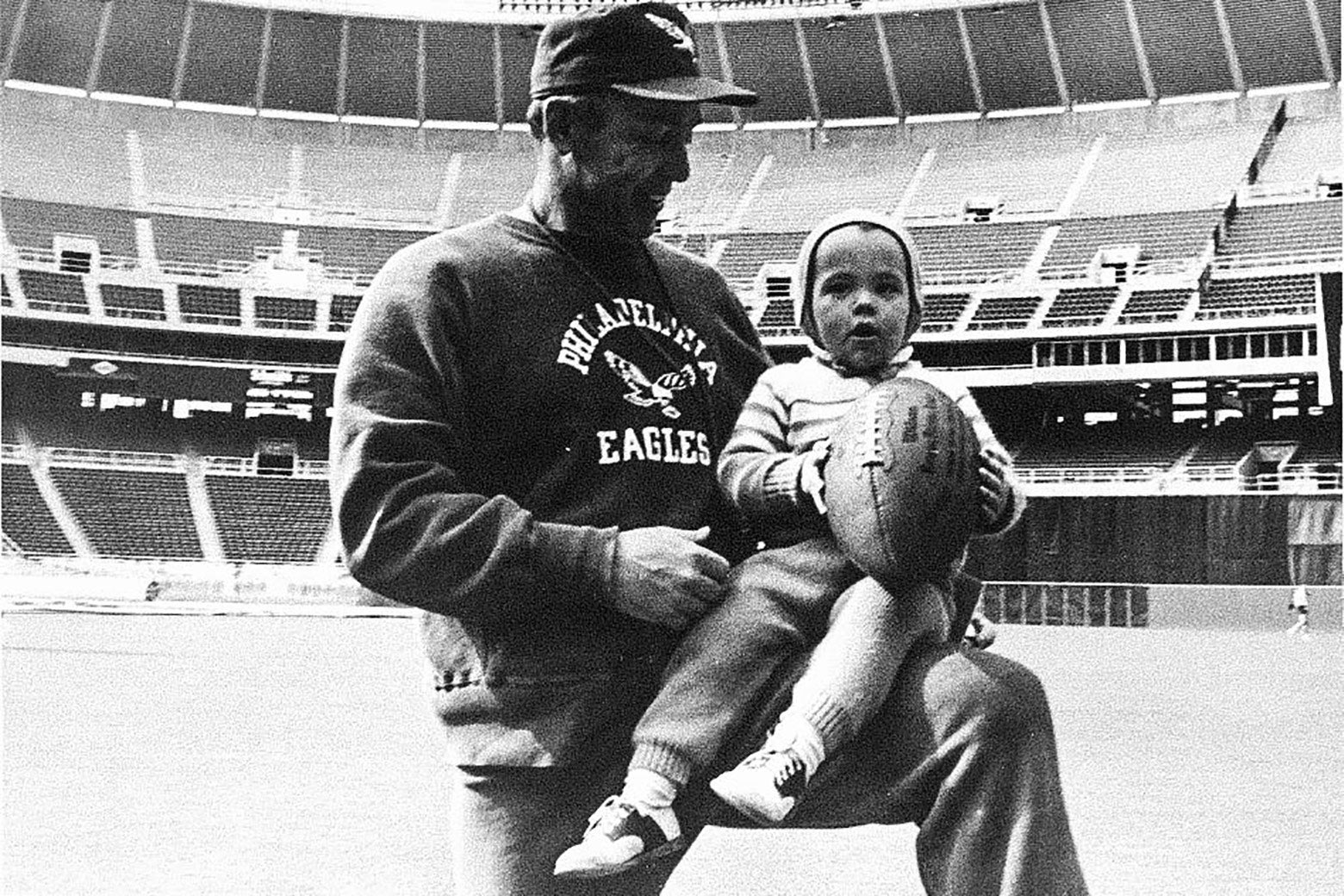 A black-and-white photo of the author as a toddler, holding a football, on the knee of his father inside a football stadium.