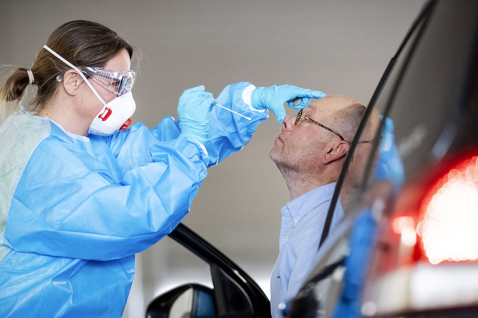 A medical worker wearing protective gear pushes up a patient's nose with one hand and lowers a long swab toward the nose with the other hand