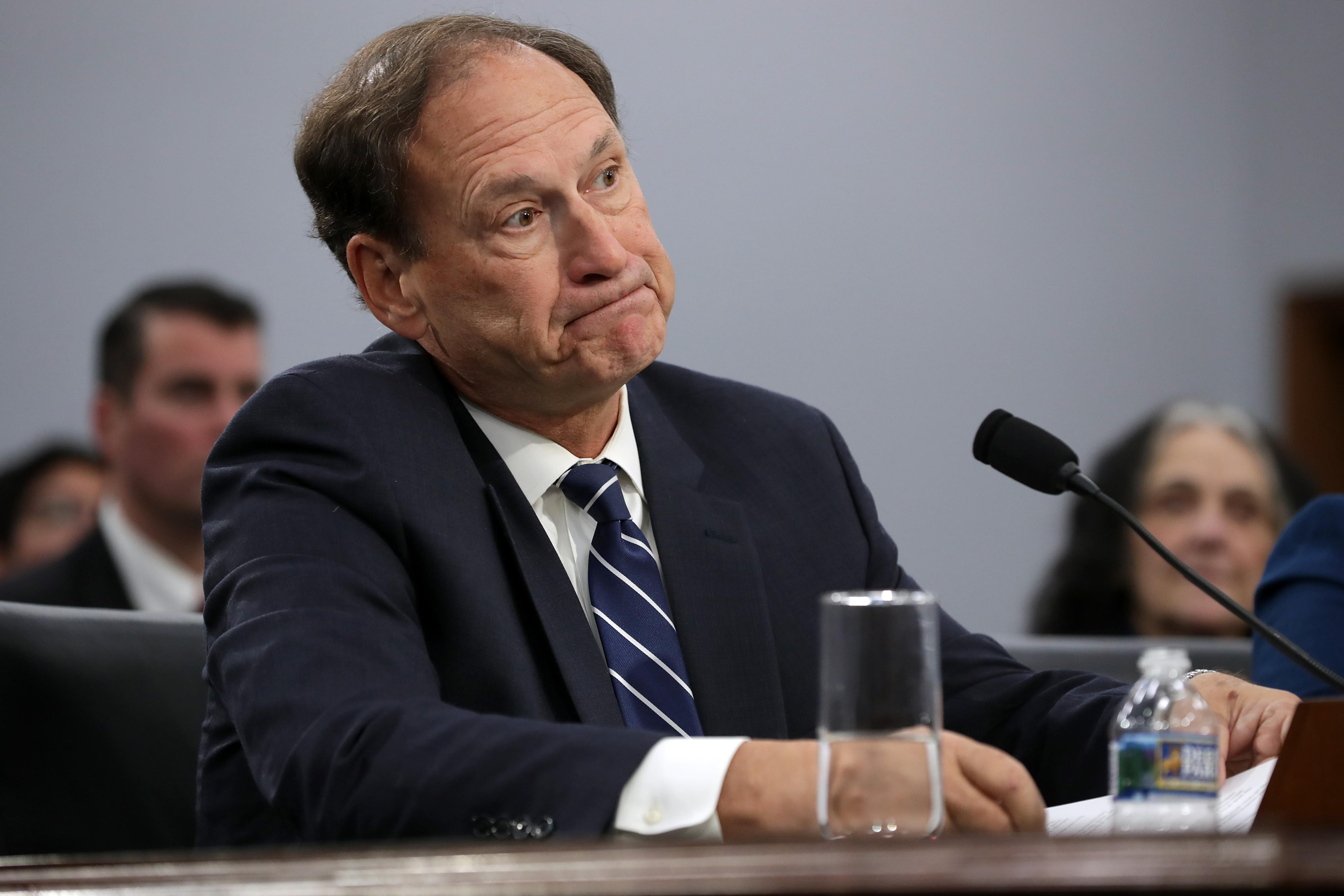 Justice Samuel Alito purses his lips while seated in front of a mic