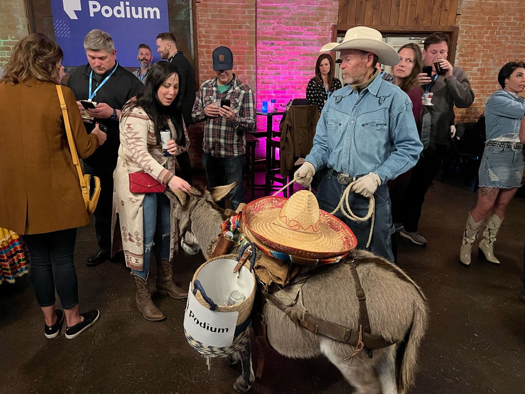 A donkey, with a sombrero on its back and loaded with baskets containing beer cans, accepts pets from a woman in the crowd.