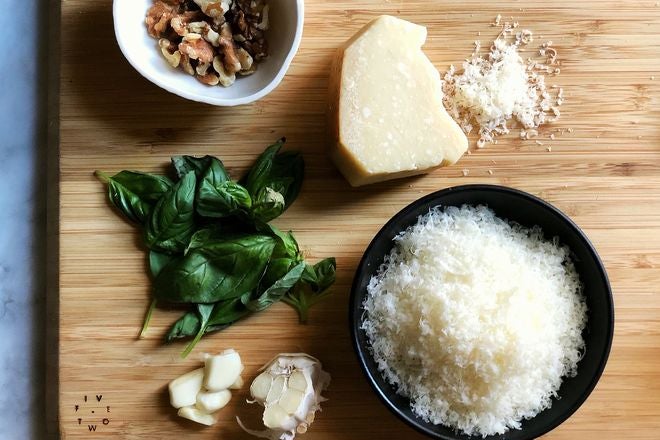 A cutting board with garlic, basil, nuts, and grated cheese.