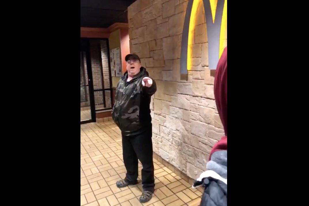 The man, wearing a camo sweatshirt, can be seen pointing a finger toward a teenager wearing a hijab. A McDonald's "M" is seen behind him.