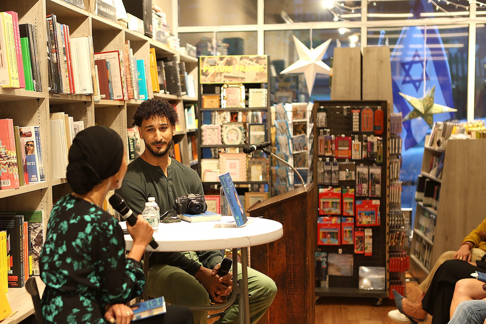 The author speaks with a moderator, Mafaz Al-Suwaidan, at his event. Through the window, we see a large Israeli flag from the protesters outside.