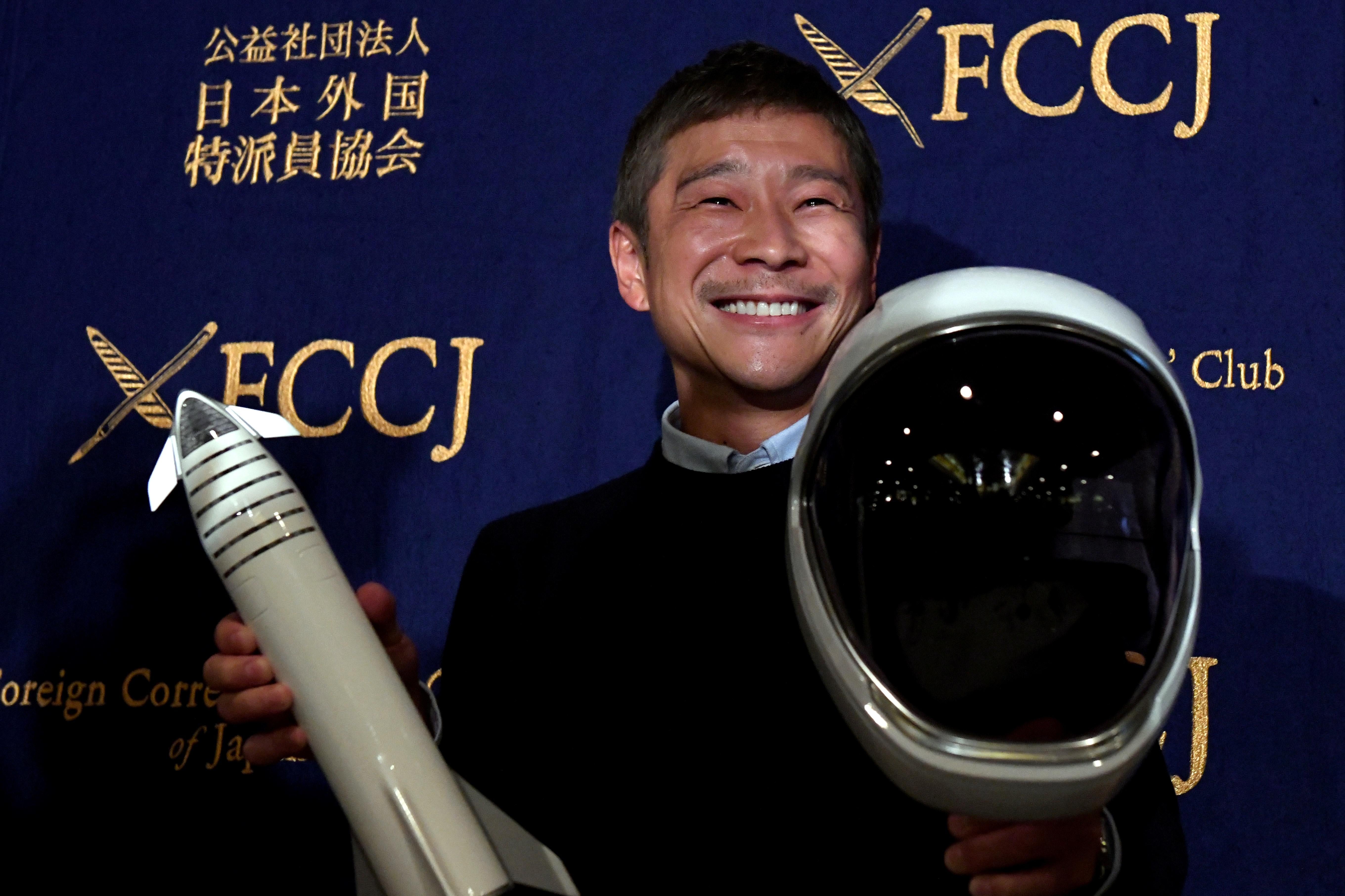 Yusaku Maezawa holds a model rocket in one hand and a space helmet in the other at a SpaceX event. He is smiling broadly and wearing a collared shirt under a sweater.