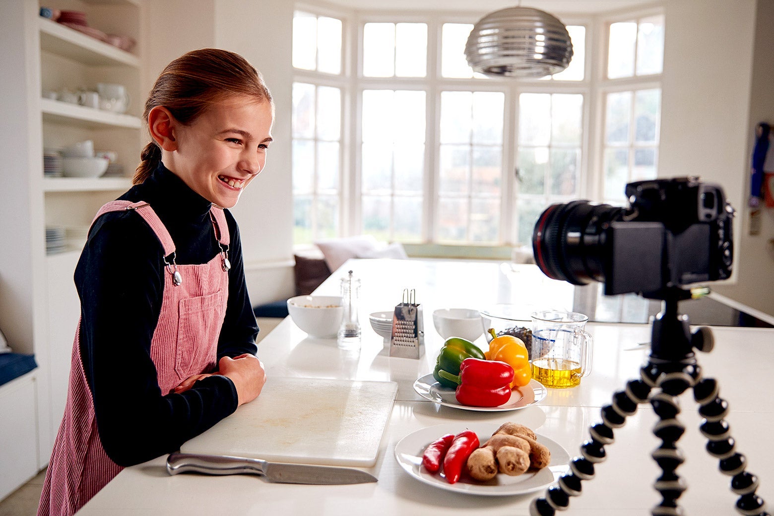 A young girl standing in a kitchen grins at a camera set up on the counter.