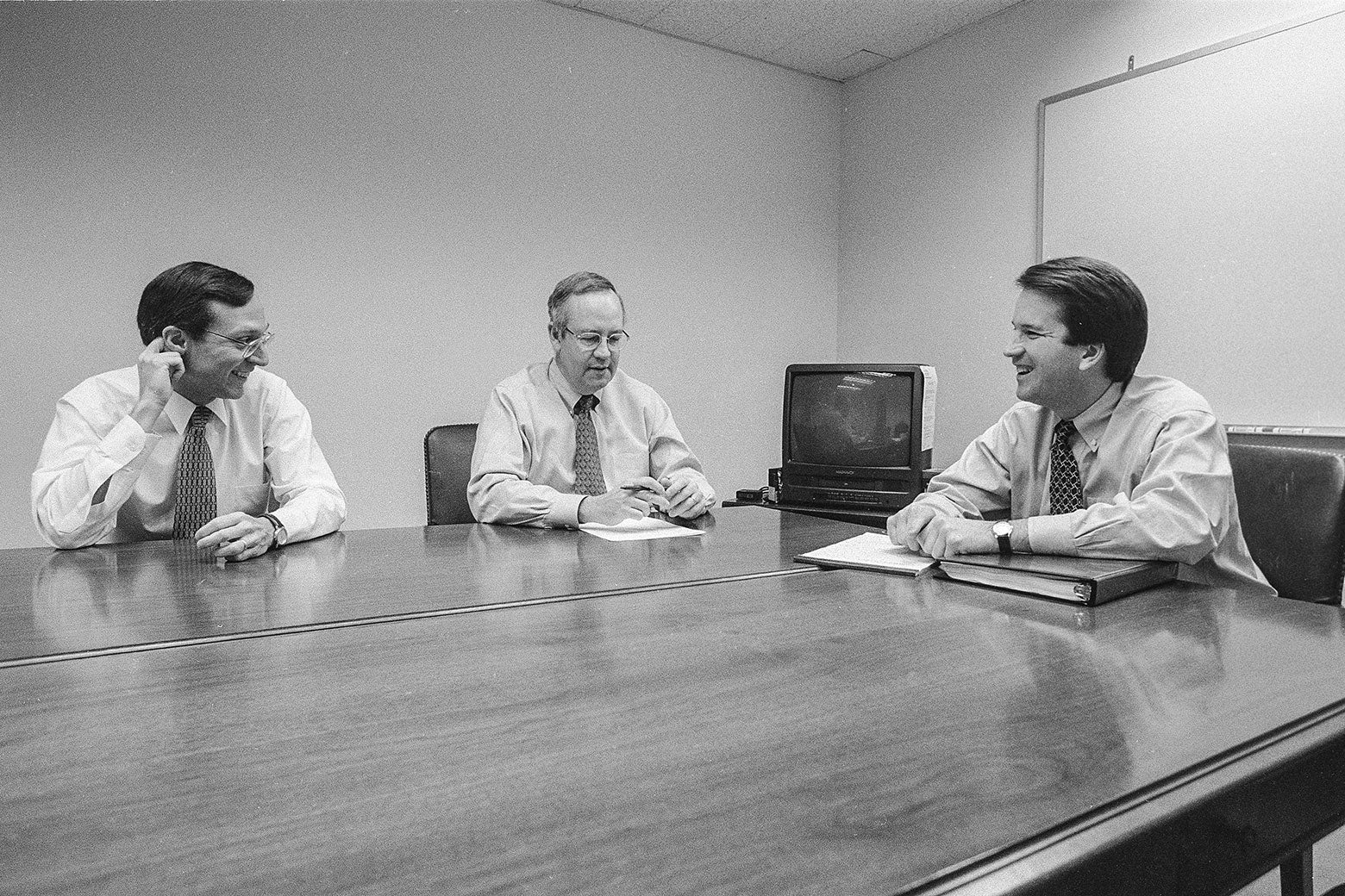 Black and white photo of Starr, Bates, and Kavanaugh sitting at a conference table in a drab room. Bates and Kavanaugh smile broadly.