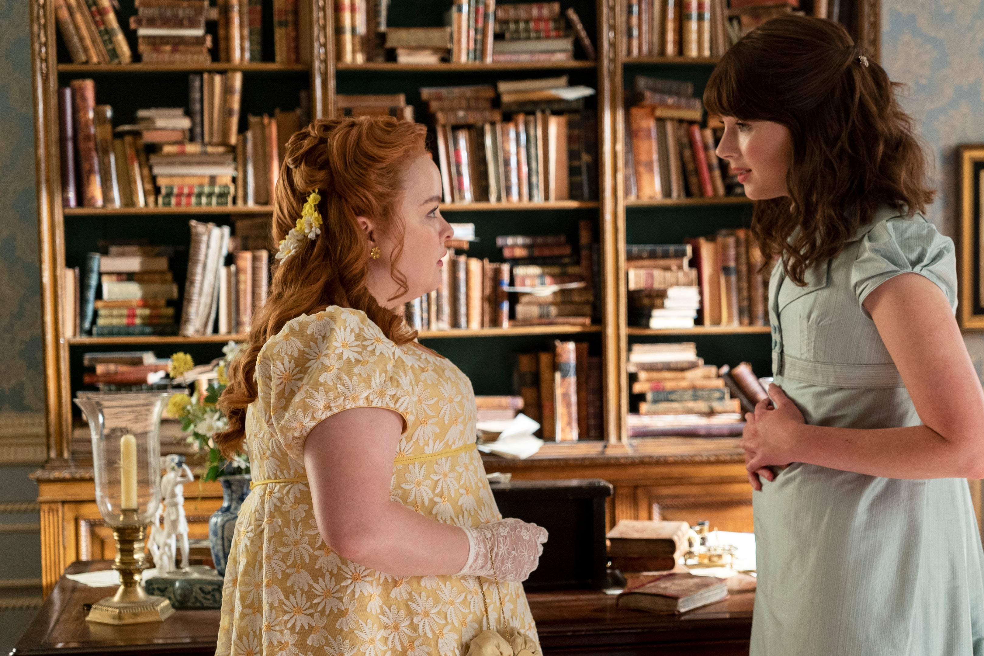 Nicola Coughlan as Penelope Featherington and Claudia Jessie as Eloise Bridgerton stand facing one another in front of a desk and bookshelf.