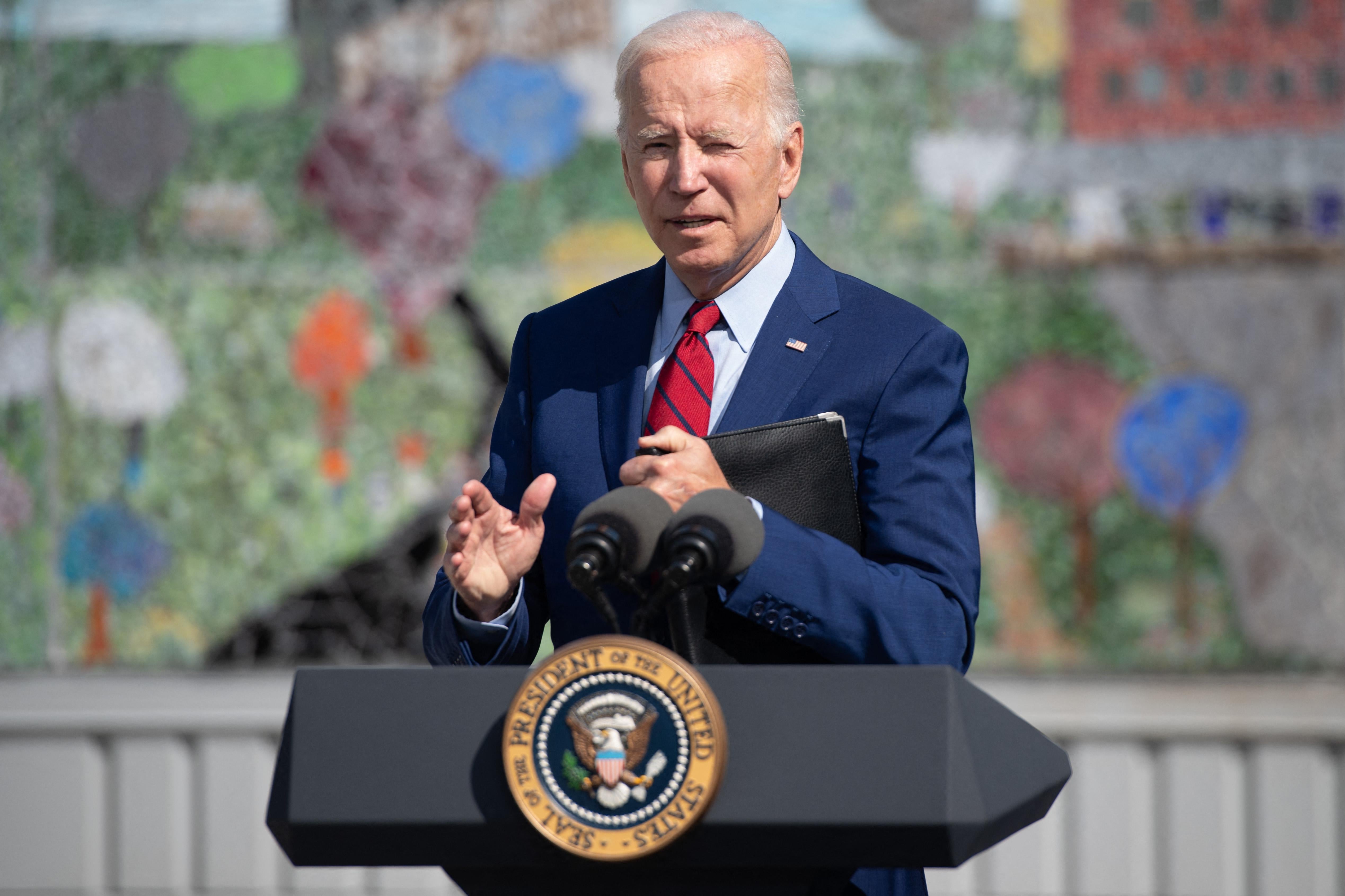 Joe Biden stands outside behind a presidential podium with a mural behind him.