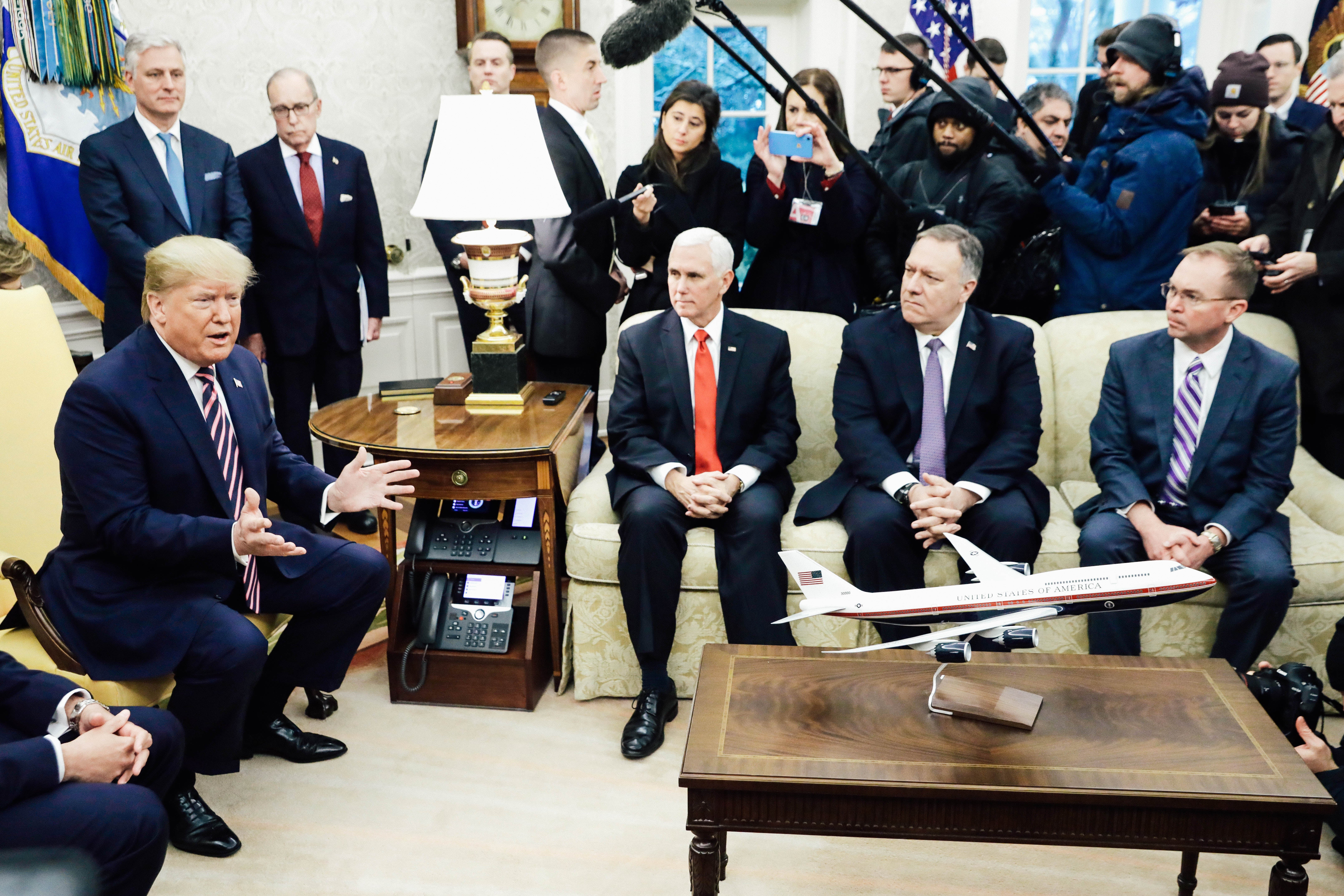 On the left, Donald Trump sits on a chair and speaks, with Robert O'Brien and Larry Kudlow standing behind. Mike Pence, Mike Pompeo, and Mick Mulvaney sit on a couch near Trump and look at him as reporters hold up microphones behind them.