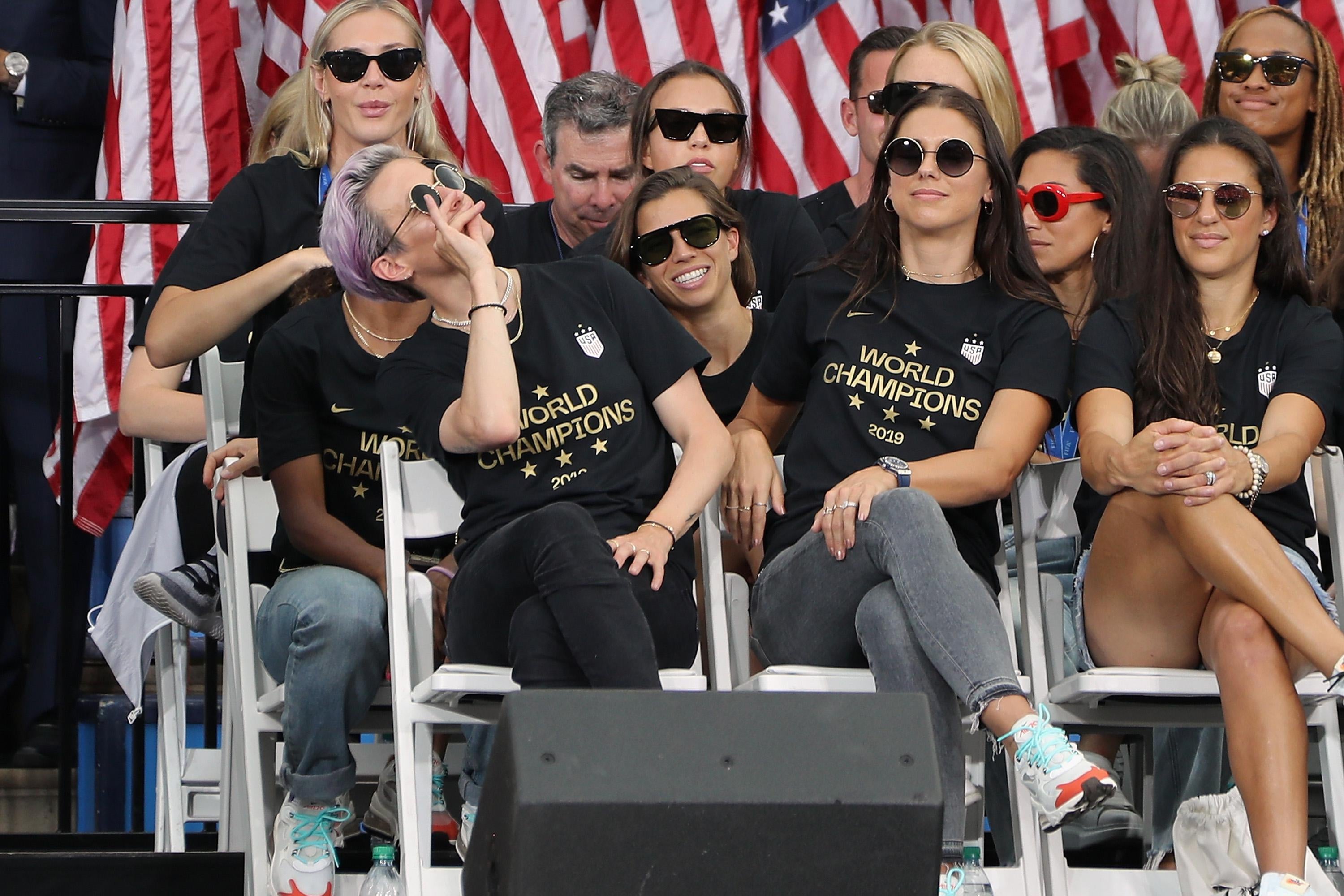 Megan Rapinoe pretends to sip tea, seated next to Alex Morgan on a stage, with several other USWNT players around them. They are all wearing sunglasses. 