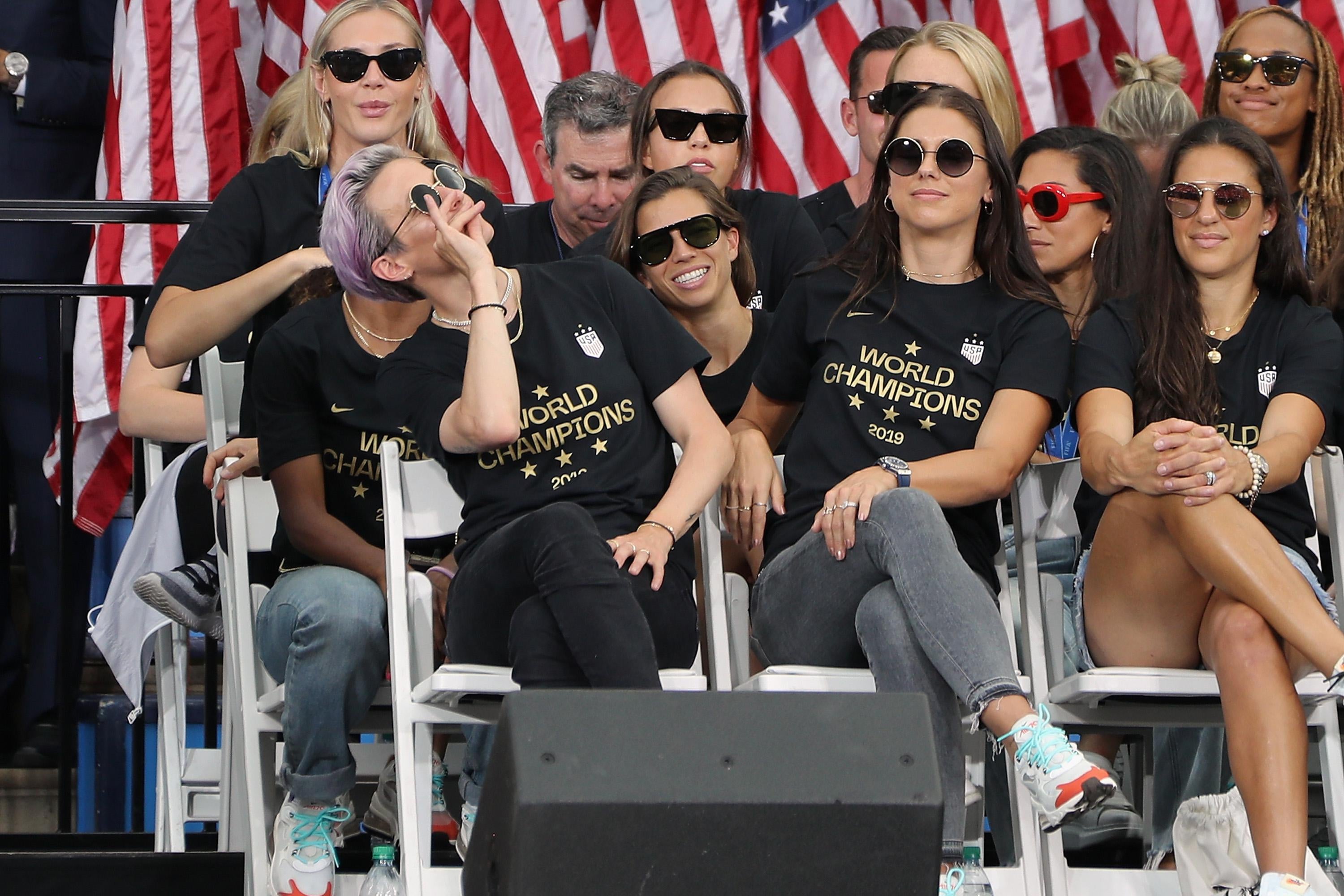 Megan Rapinoe pretends to sip tea, seated next to Alex Morgan on a stage, with several other USWNT players around them. They are all wearing sunglasses.
