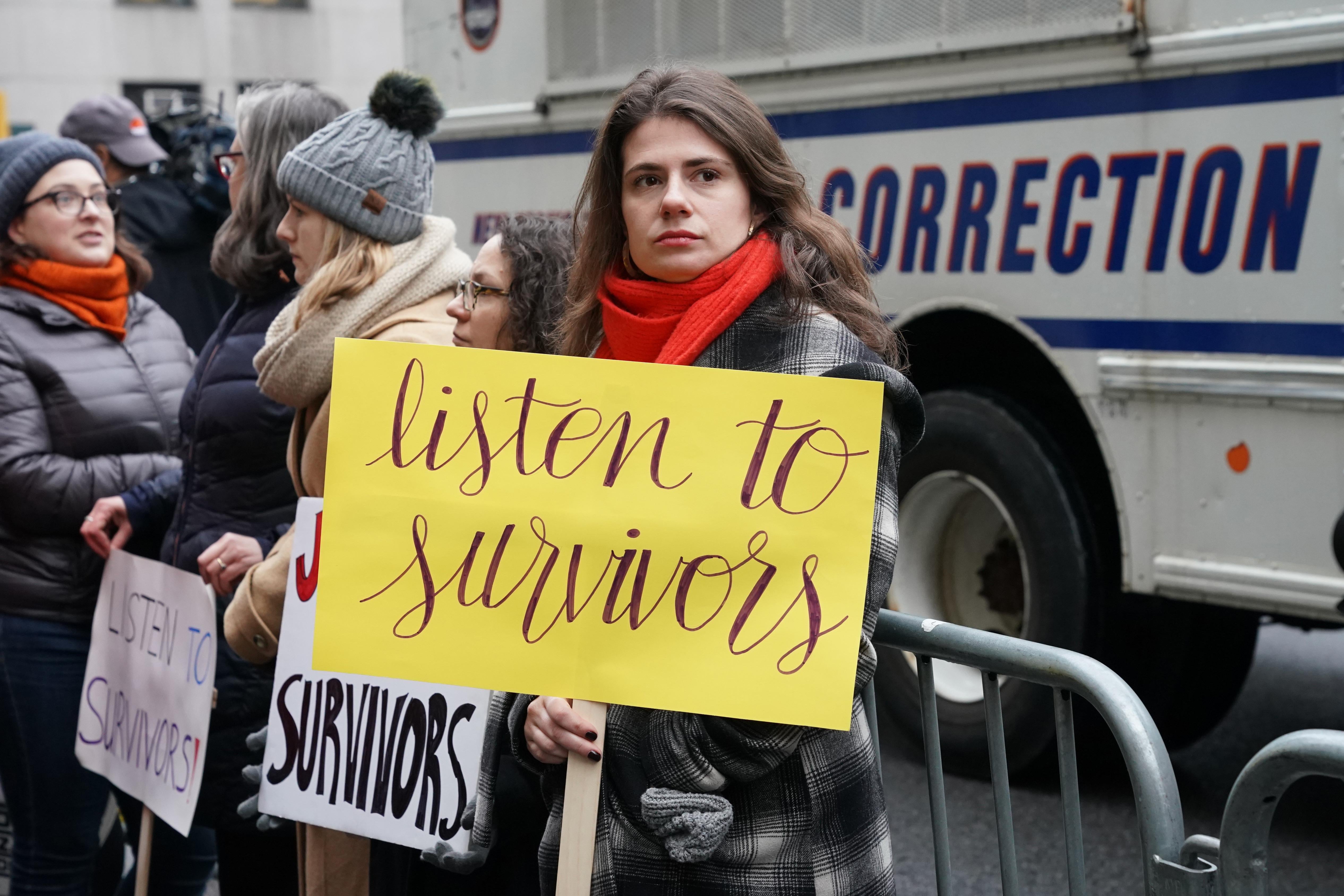 A woman holds a sign that says "listen to survivors" standing outside the courthouse on the first day of Harvey Weinstein's trial. 