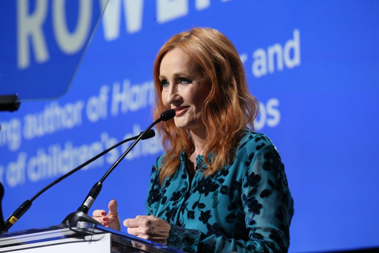 J.K. Rowling stands behind a podium wearing a blue dress.