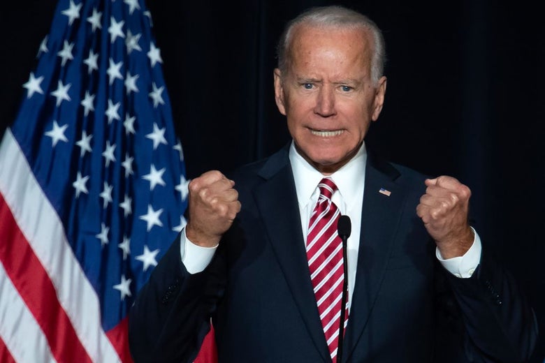 Biden clutches his fists dramatically while speaking against a dark blue backdrop and next to an American flag.