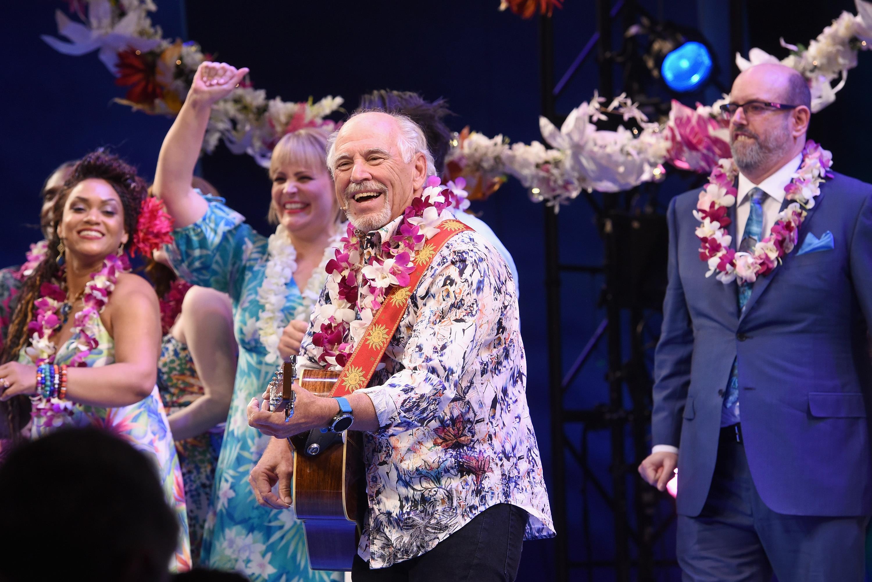 He smiles wide on a stage garlanded with leis, a lei over his shoulder,s white hair on his head, and little golden suns on the strap of his acoustic guitar.