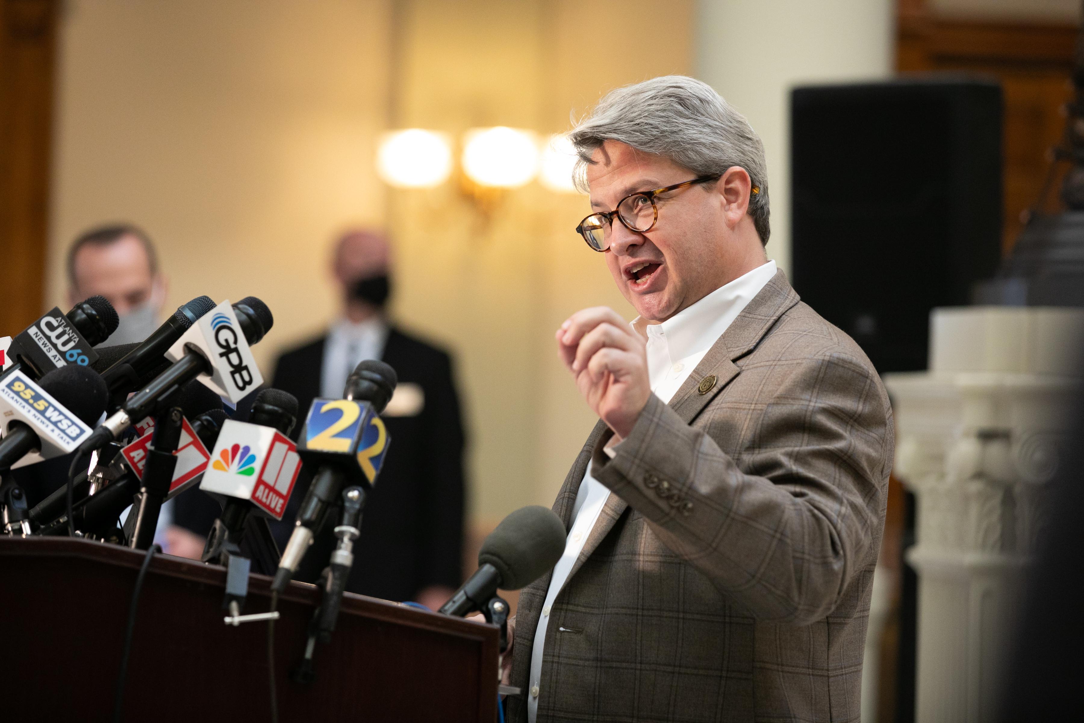 Gabriel Sterling, wearing a tan suit, speaks into a number of microphones.