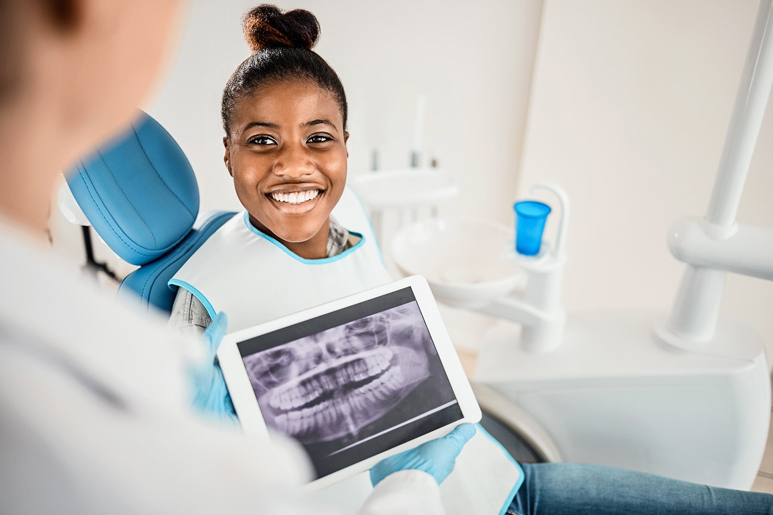 A young woman smiles while seated in a dentist's chair as a dental professional looks at her x-rays.